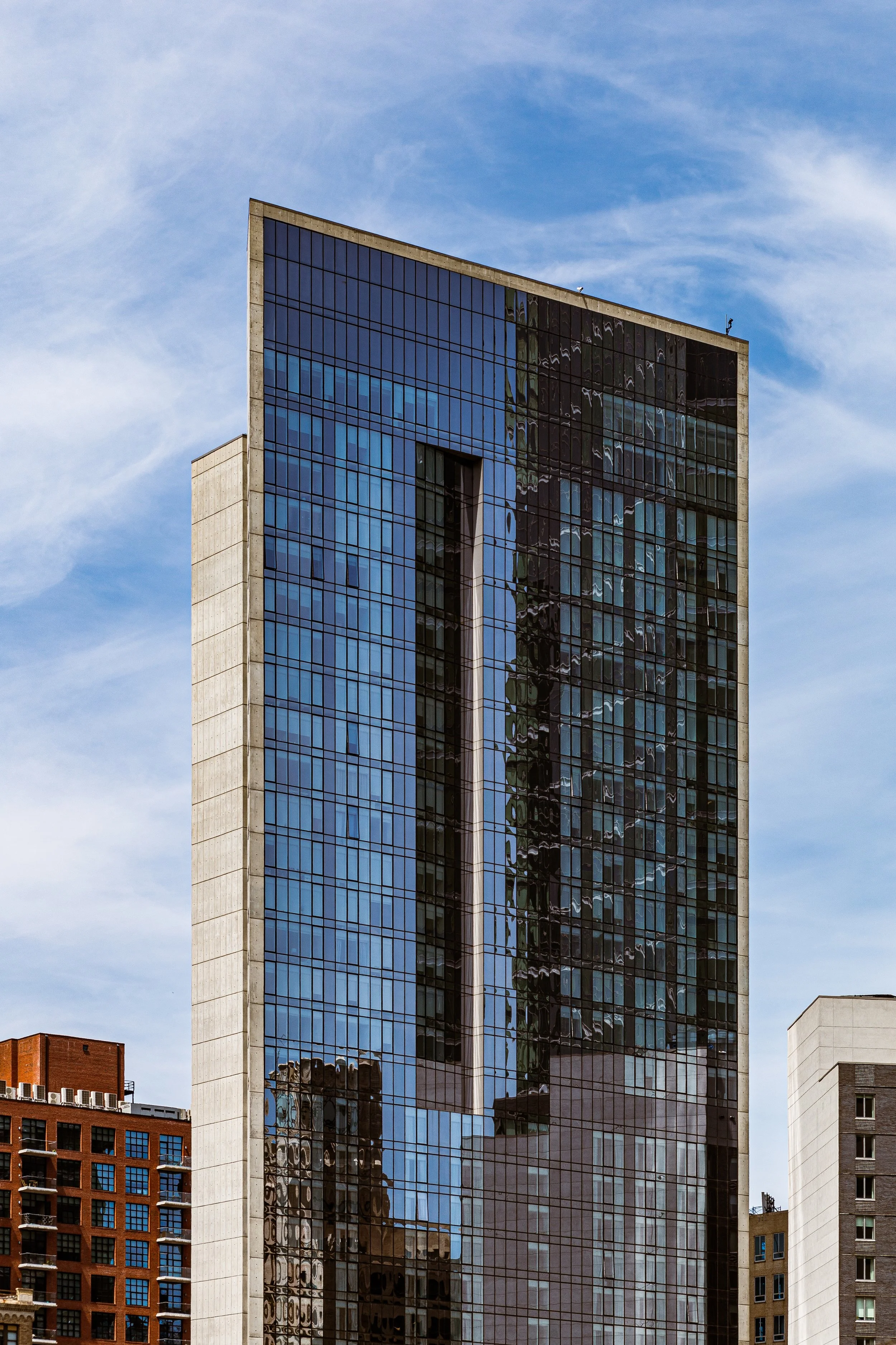Tall modern skyscraper with dark reflective glass windows reflecting neighboring buildings and sky, against a blue sky with wispy clouds.