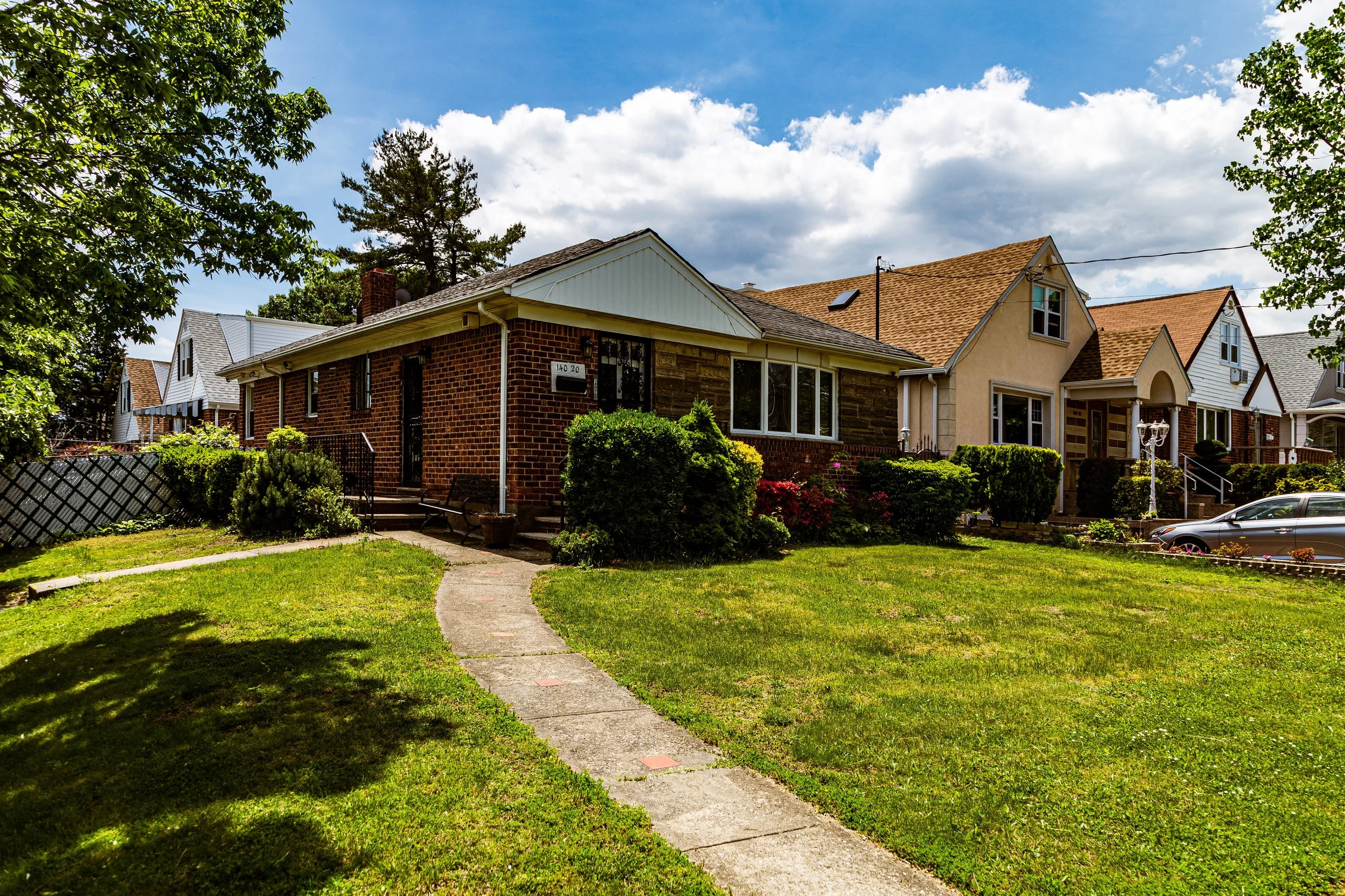 View of a suburban neighborhood with brick and stucco houses, green lawns, and a winding walkway on a sunny day with partly cloudy sky.