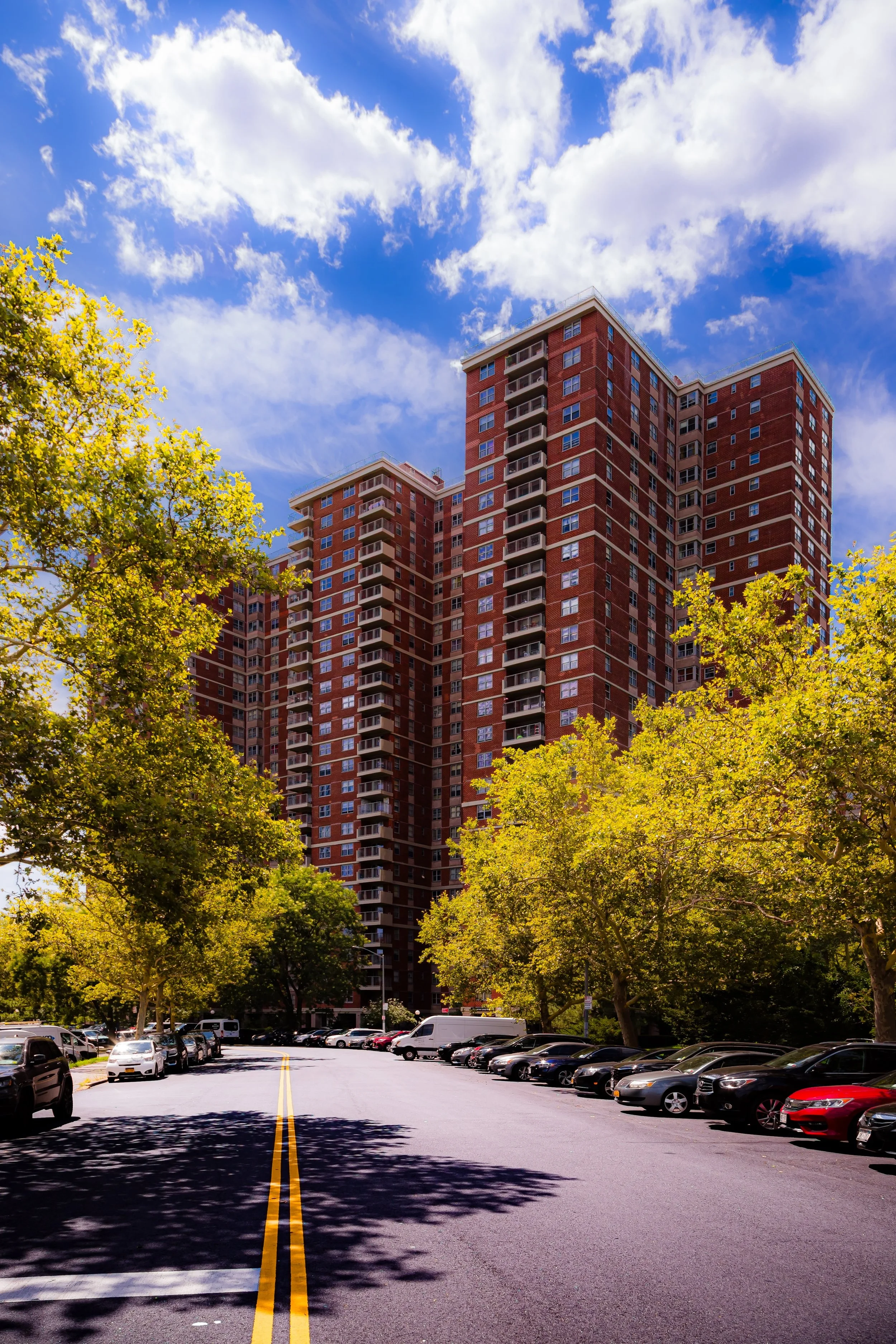 Tall red-brick apartment building with multiple balconies surrounded by green trees and parked cars on a sunny day with partly cloudy skies.