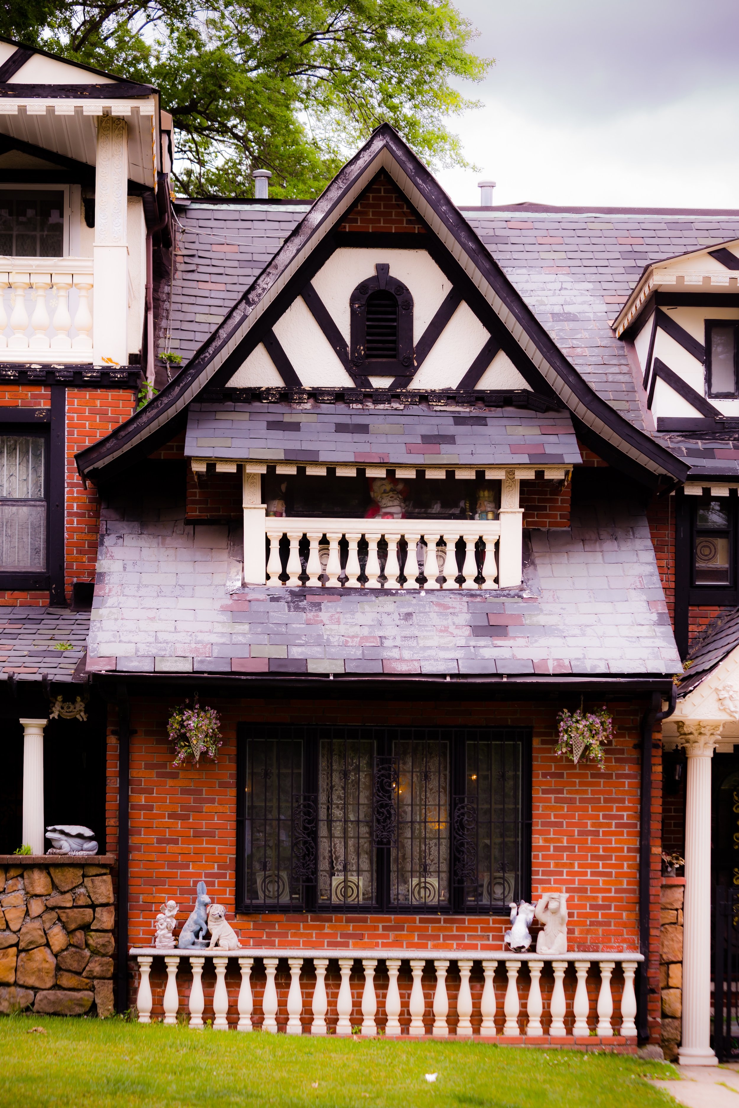 A two-story brick house with a steep gabled roof, decorative black and white woodwork, and a front porch with white balustrades. The house features a large window with iron bars and several animal statues and hanging flower baskets in front.