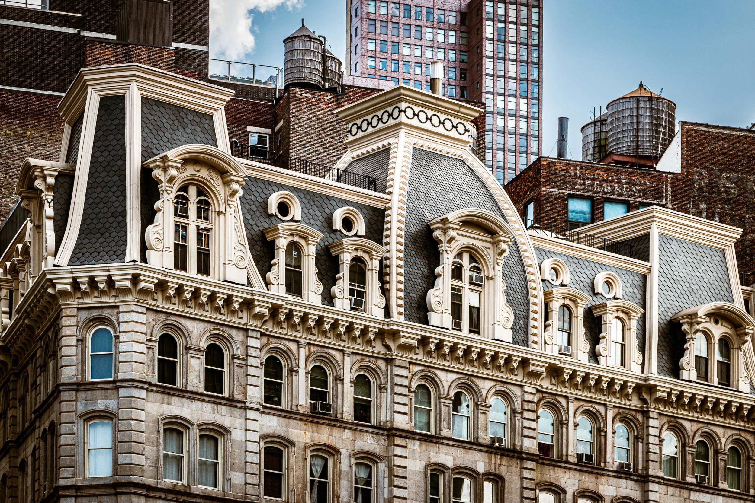 Close-up view of an ornate, historic building with detailed stonework, dormer windows, decorative trim, and a contrasting dark shingle roof, set against taller modern skyscrapers in the background.