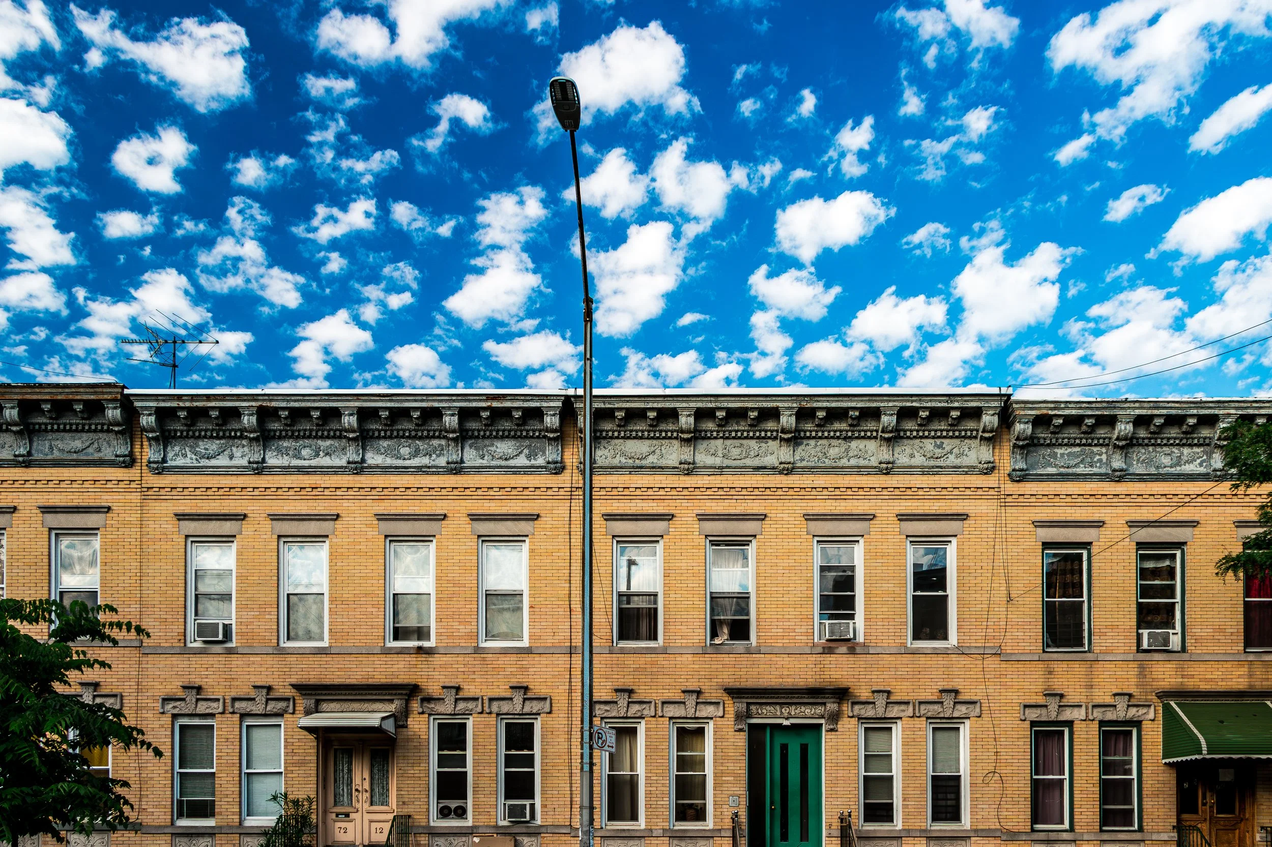 A row of brick apartment buildings with multiple windows and air conditioning units, under a bright blue sky with scattered clouds, and a streetlamp in the foreground.