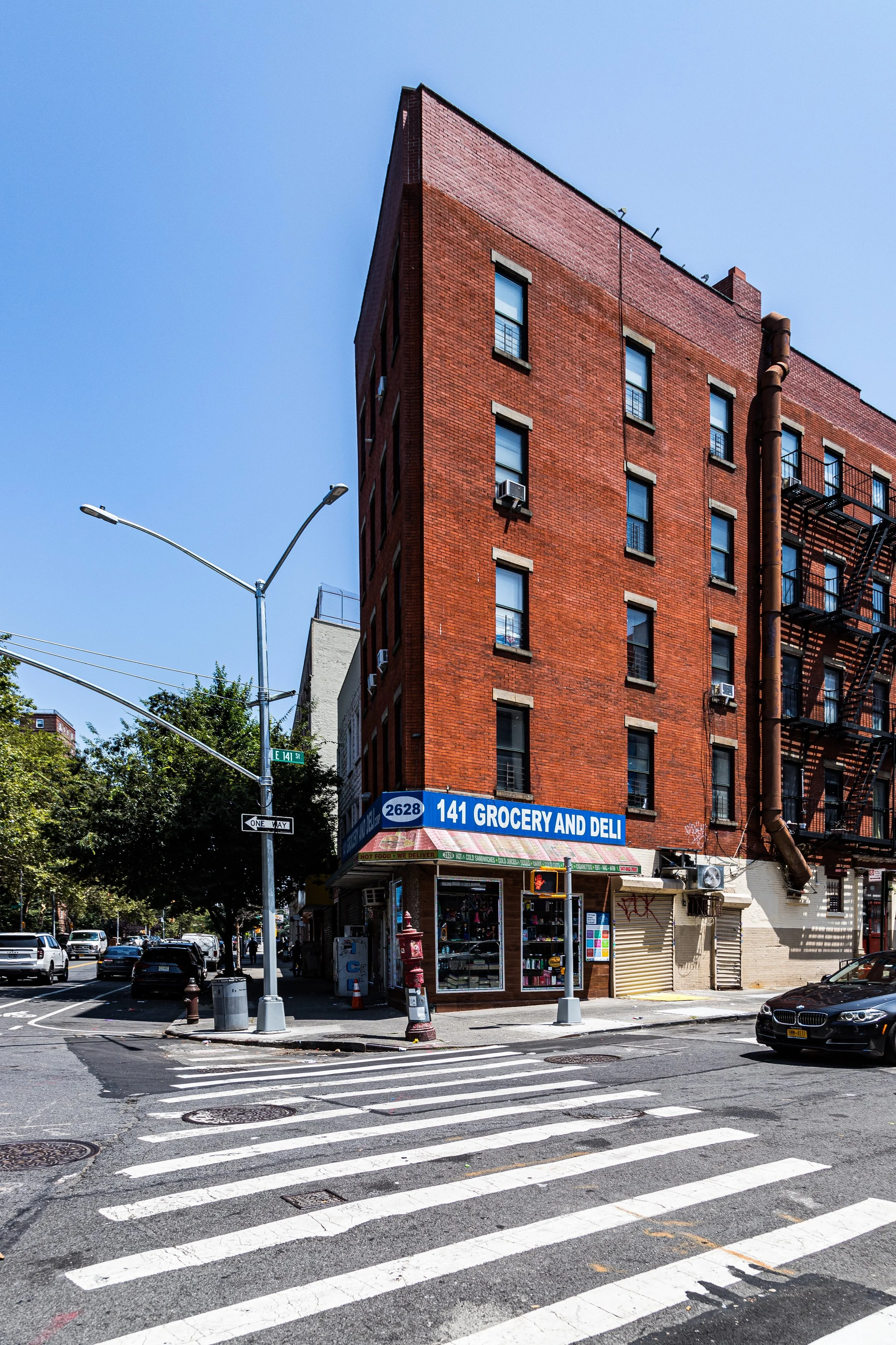 A corner of a city street showing a tall red brick building with a grocery and deli on the ground floor, and a zebra crossing in the foreground.