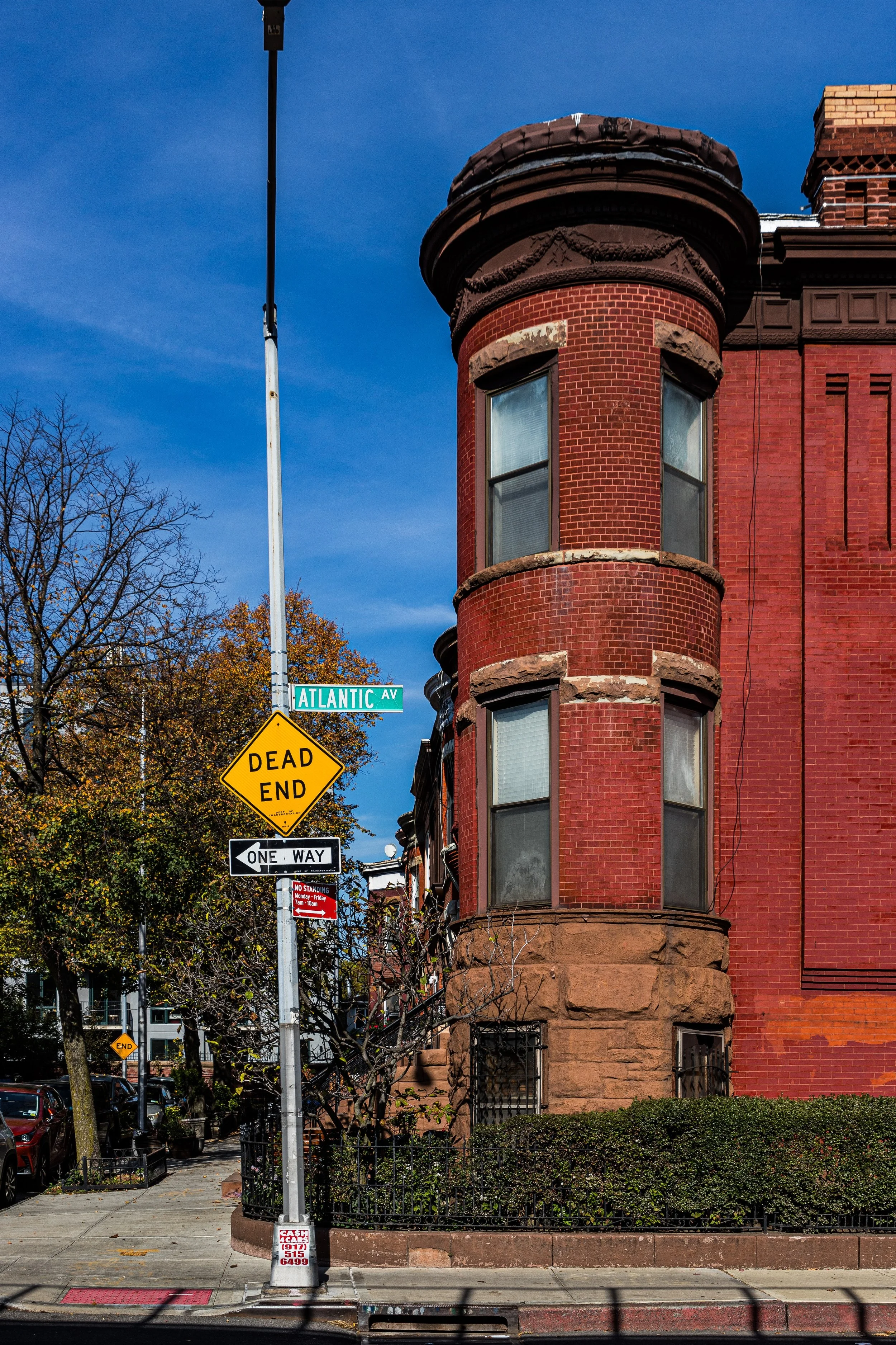 A corner building with a rounded tower, made of red brick with stone accents, on Atlantic Avenue. There are street signs for Atlantic Avenue and a dead end sign, along with a one-way sign and other street signs. The building has multiple windows and a small garden with bushes in front.