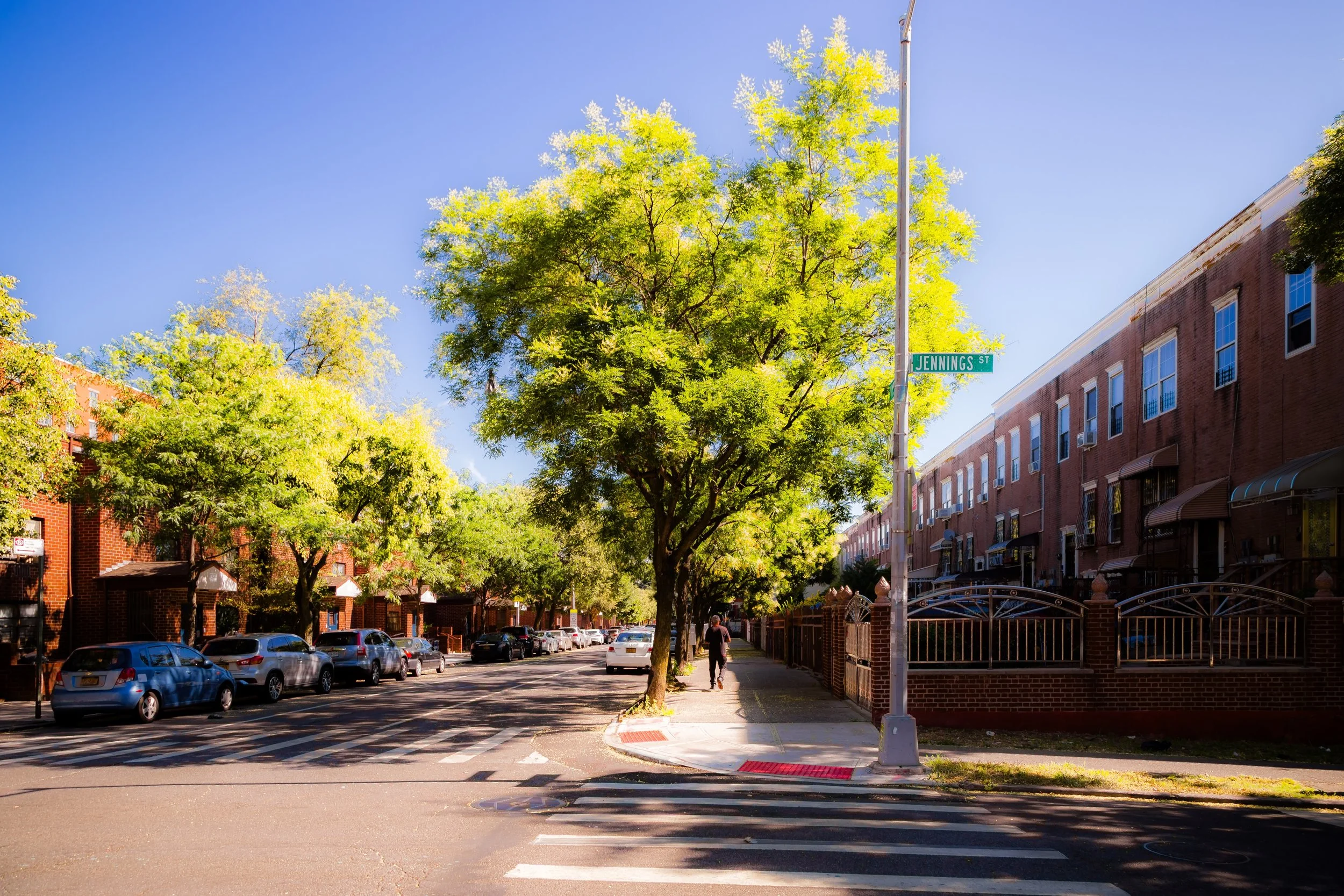 Street view of Jennings Street with parked cars on the left and brick townhouses on the right, green trees lining the sidewalk, and a person walking away on the sidewalk on a sunny day.