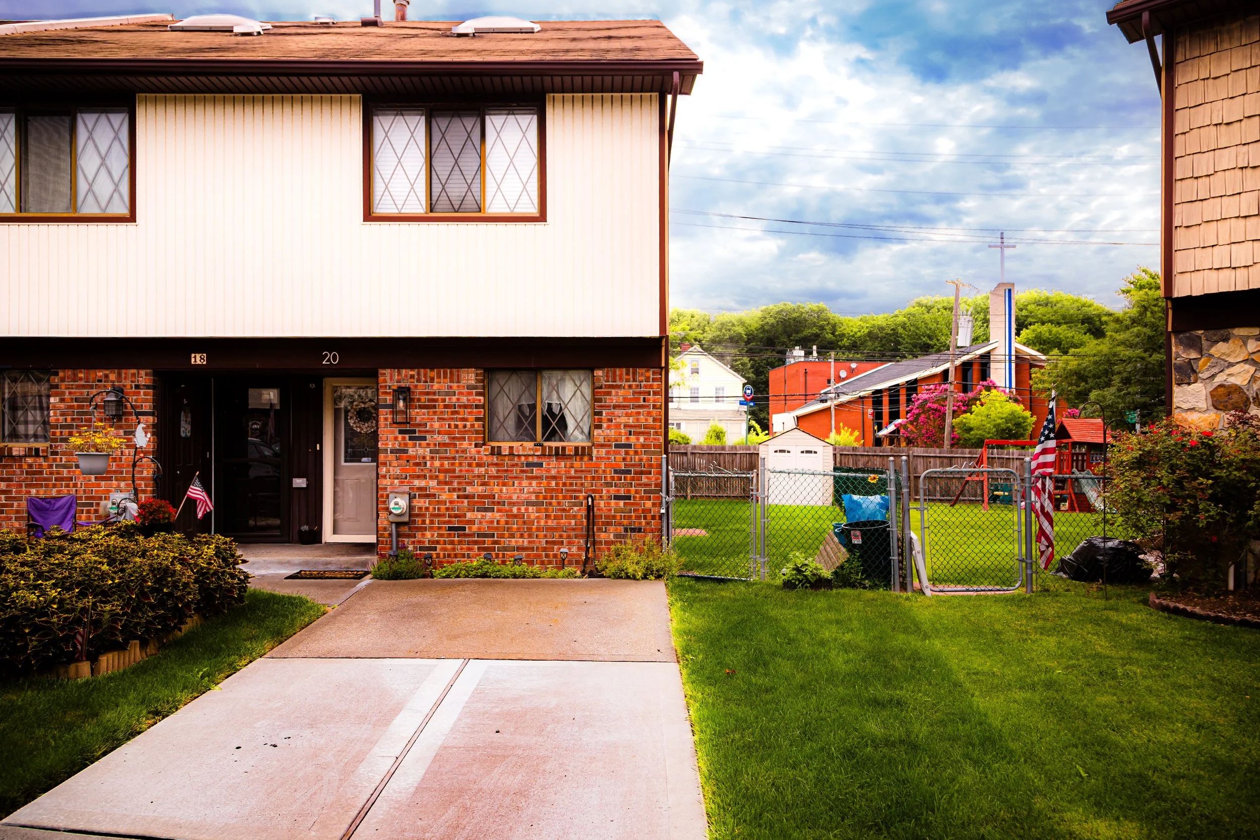 A two-story house with a brick lower level and a white upper level, a driveway, a lawn with bushes, a chain-link fence enclosing a backyard with a small shed, trash cans, bright flowers, and neighboring houses with trees in the background under a partly cloudy sky.