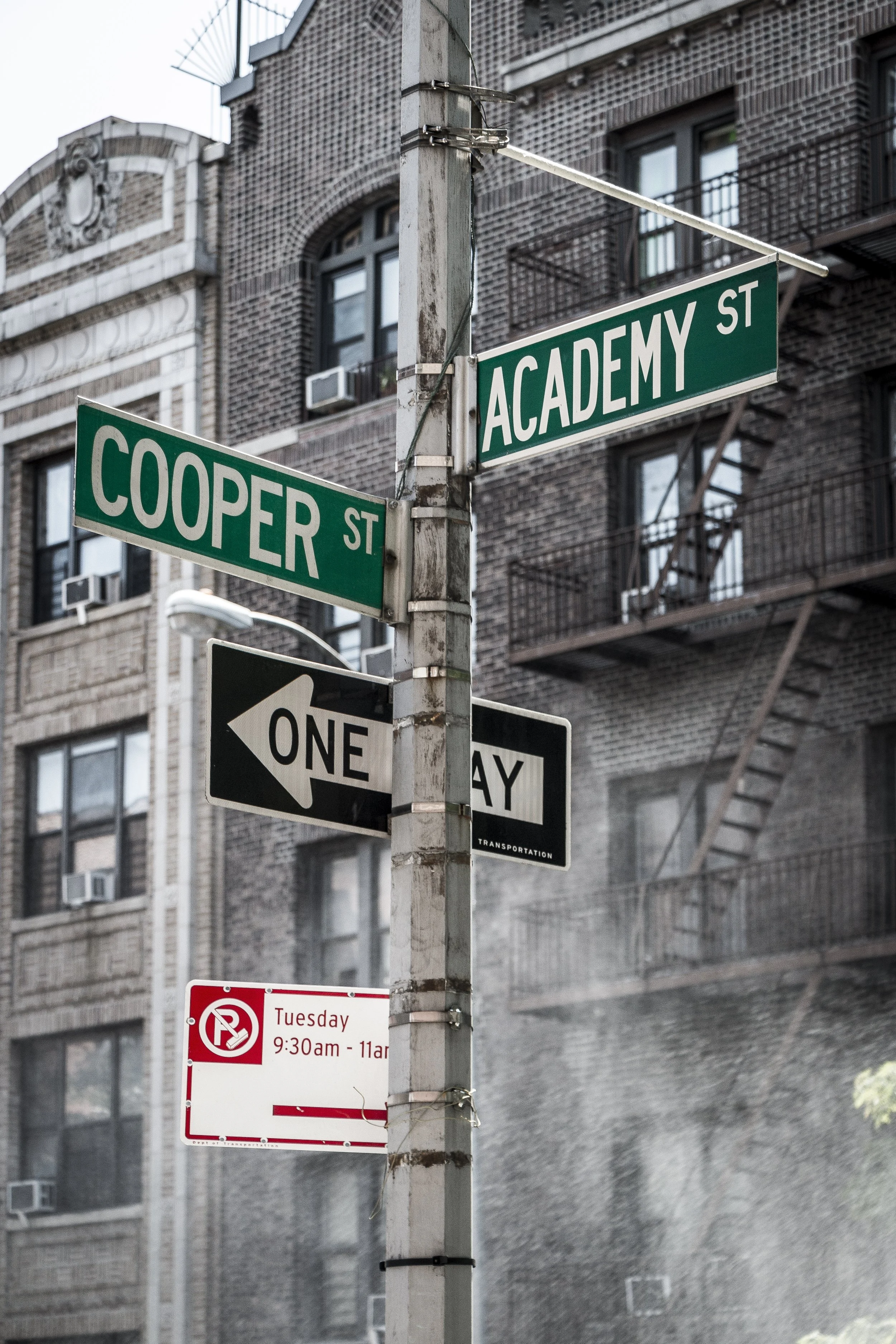Street signs at the intersection of Cooper Street and Academy Street, with a one-way sign pointing left and a no-parking sign indicating restrictions on Tuesday from 9:30 am to 11 am. In the background, there are brick apartment buildings with fire escapes.