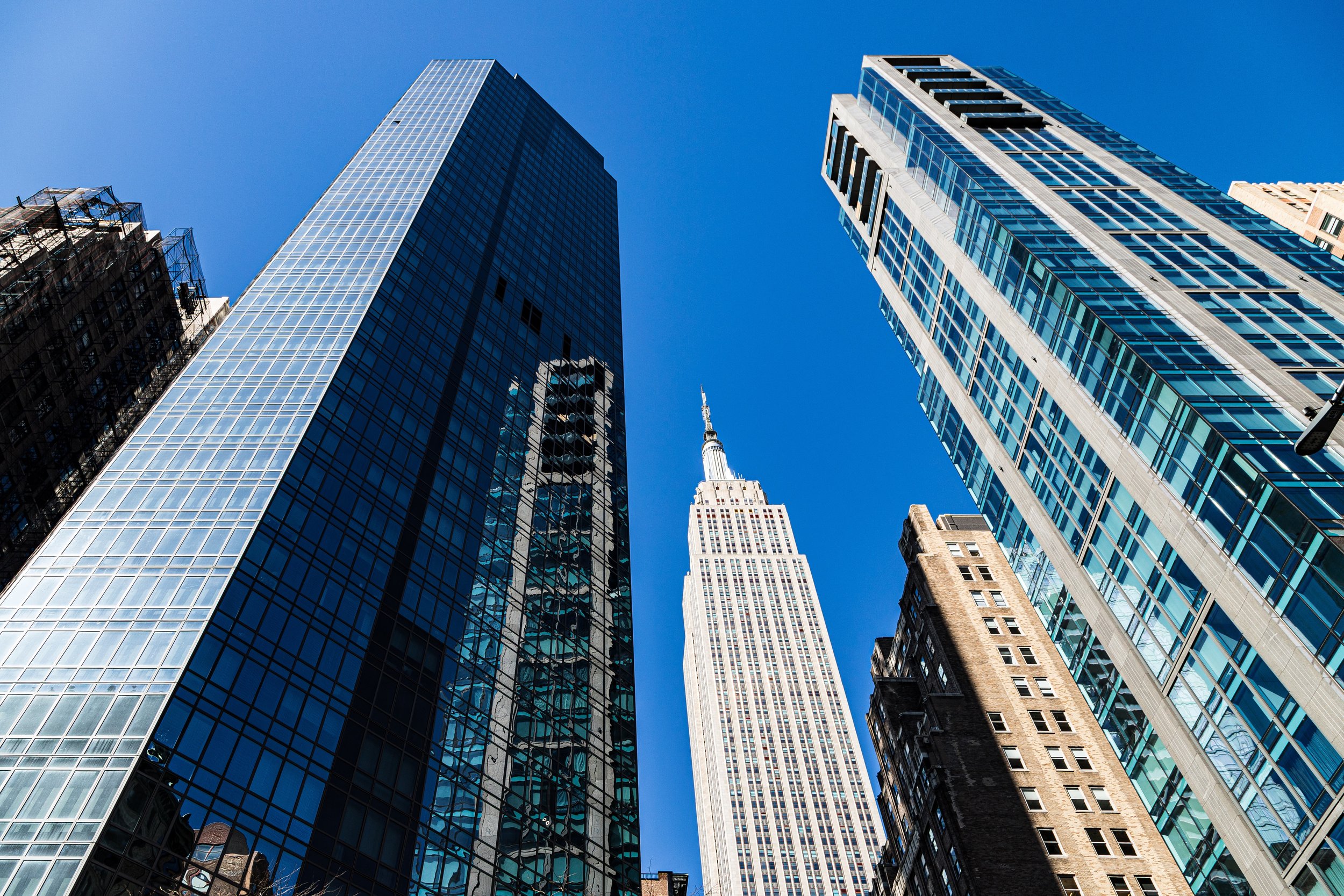 Looking up at tall skyscrapers and the Empire State Building against a clear blue sky in New York City.