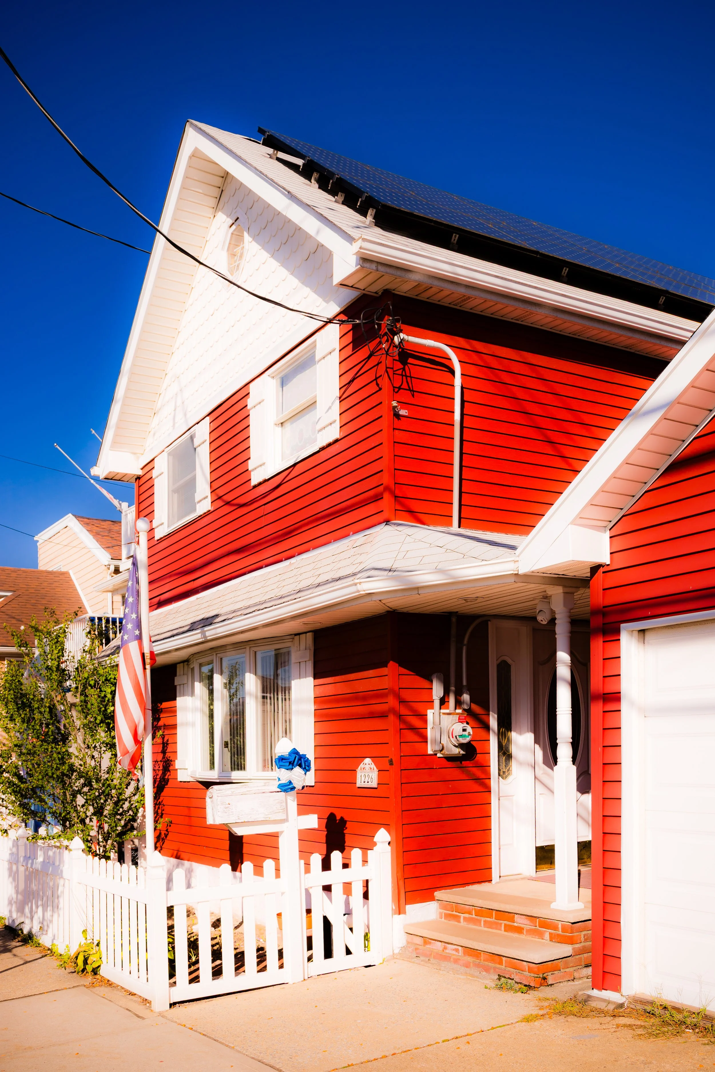 A two-story house painted red with white trim, featuring a white front porch and steps, solar panels on the roof, and an American flag in front. The house has a white picket fence and a mailbox.