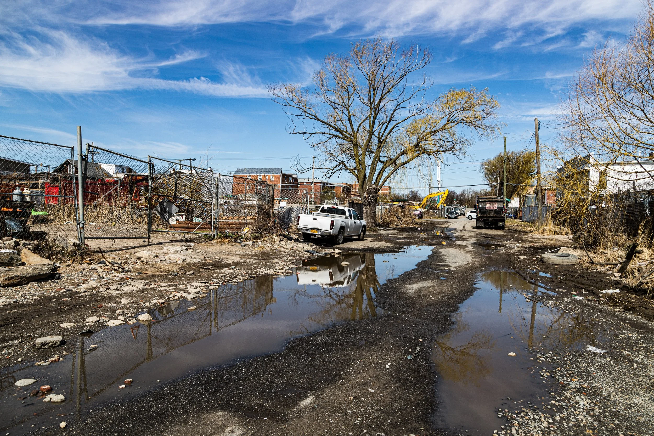 A dirt road with puddles and debris, a large tree, a white pickup truck, and construction equipment in a semi-urban area under a blue sky.