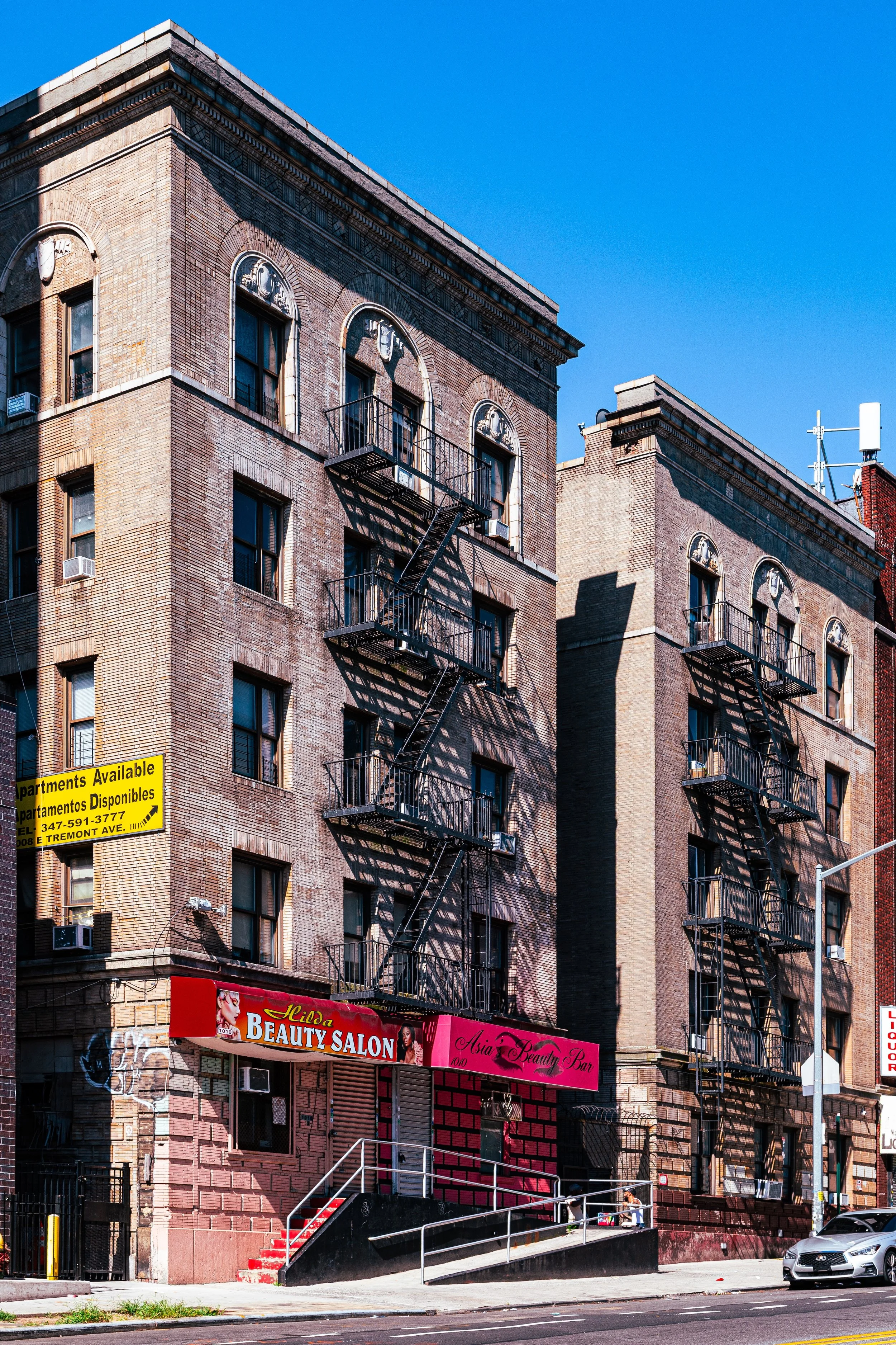 Two older brick buildings with metal fire escapes on the front, a beauty salon and a beauty parlor on the ground floor, and a yellow sign advertising available apartments. A car is parked on the street in front.