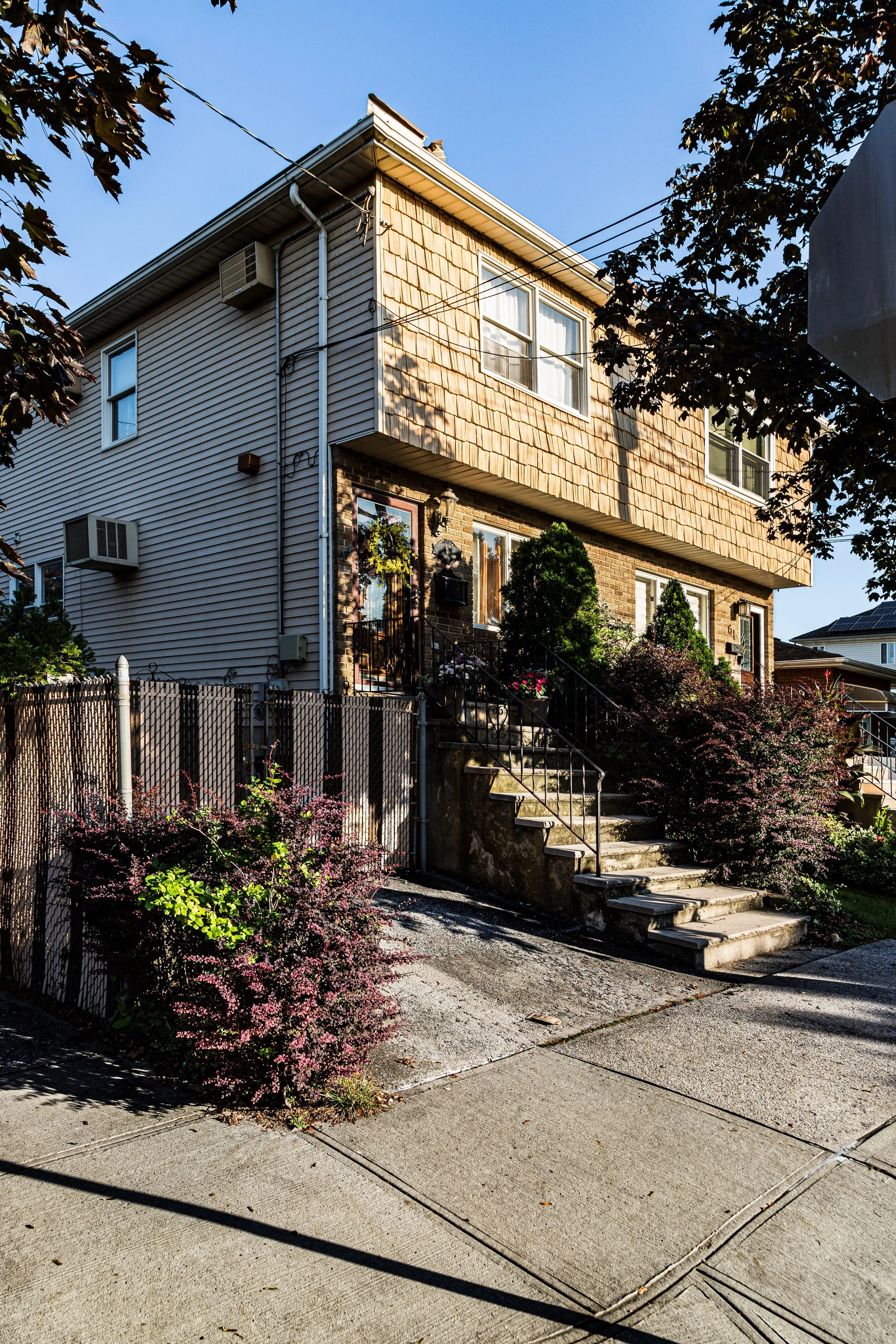 A two-story house with a brick and siding exterior, front stairs, lush shrubs, and a fence, under a clear blue sky.