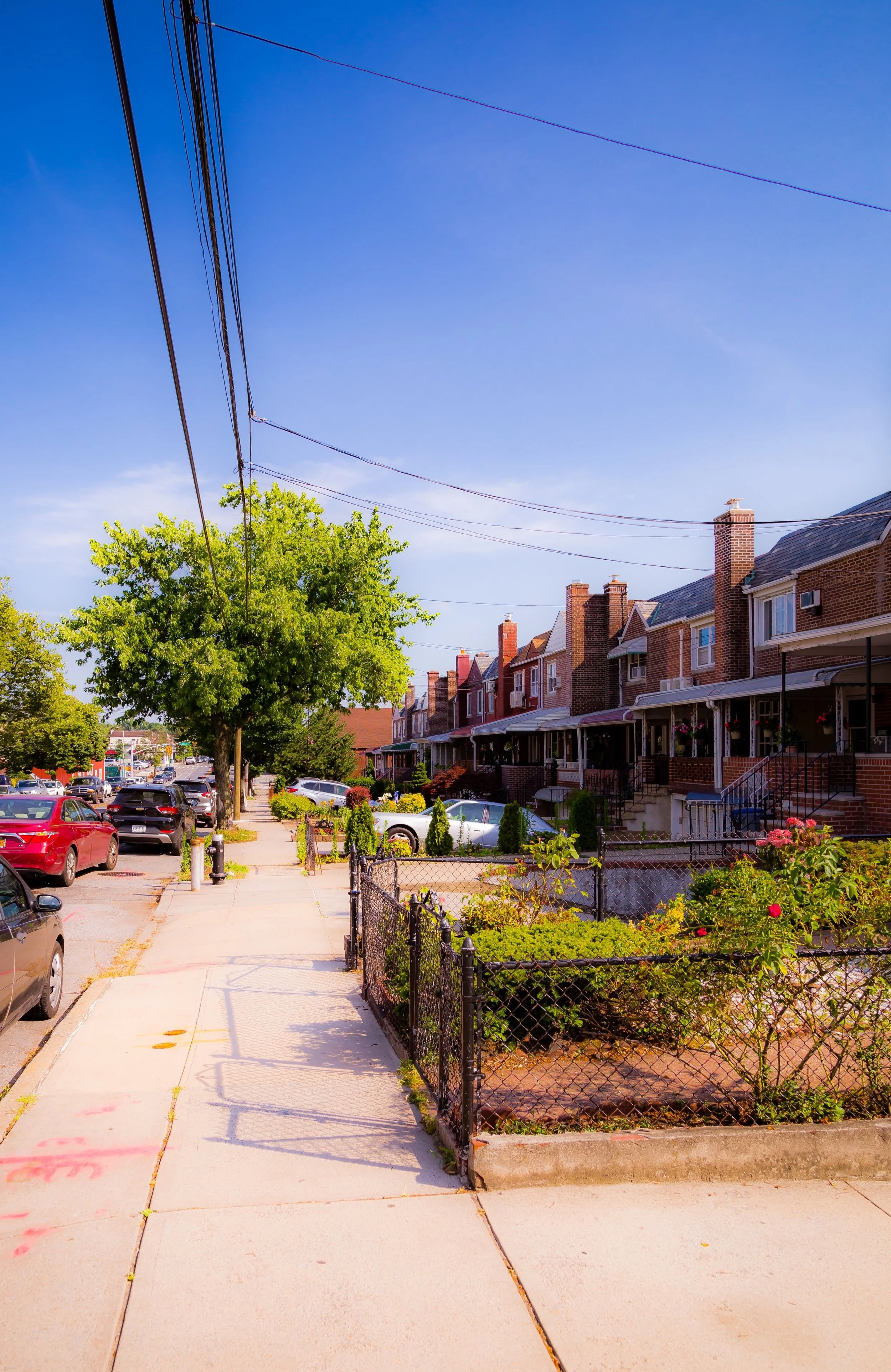 A sunny neighborhood street with parked cars on the side, a row of brick houses with porches, green trees, and a clear blue sky.