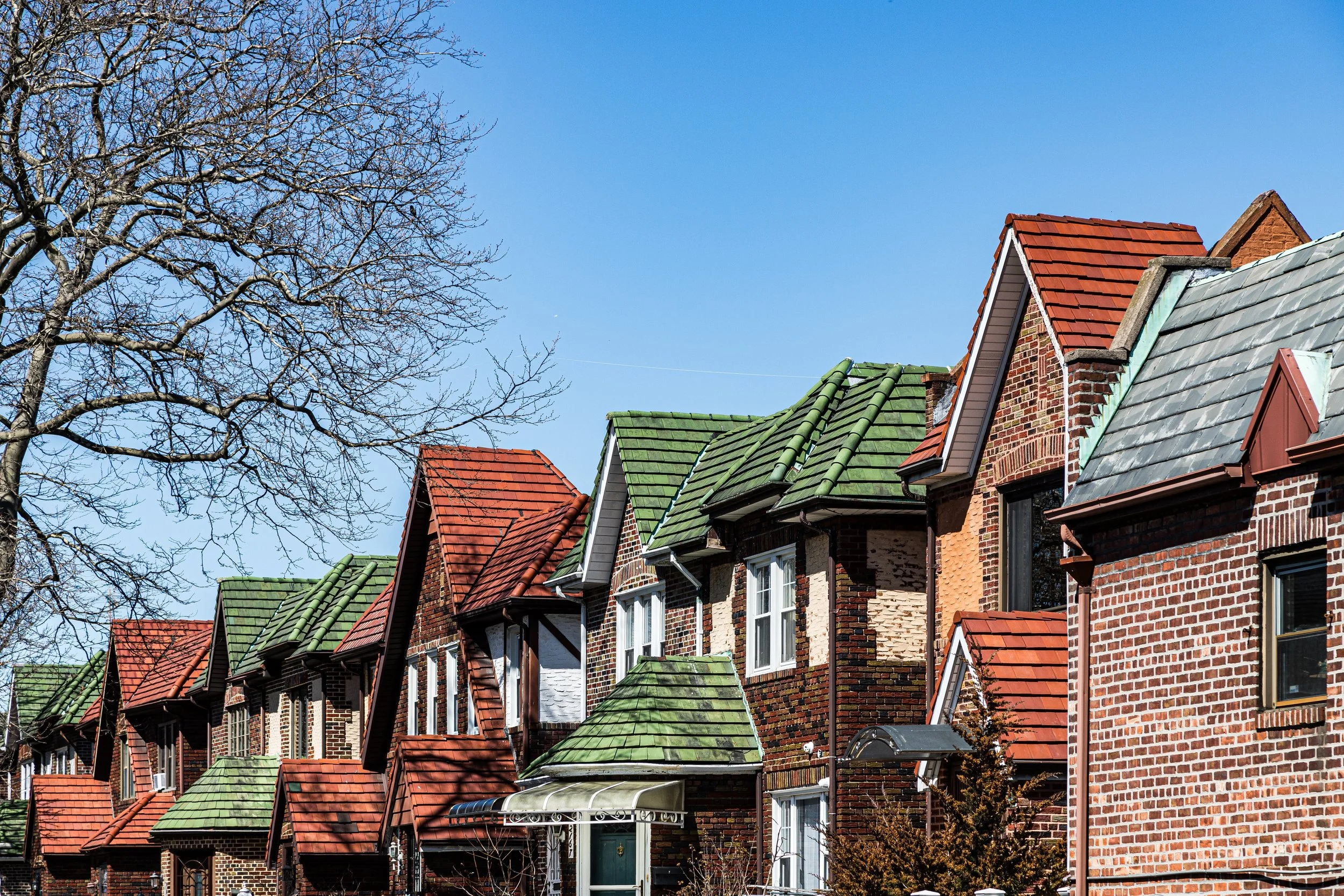 Row of brick houses with colorful tiled roofs under a clear blue sky, with a leafless tree in the foreground.