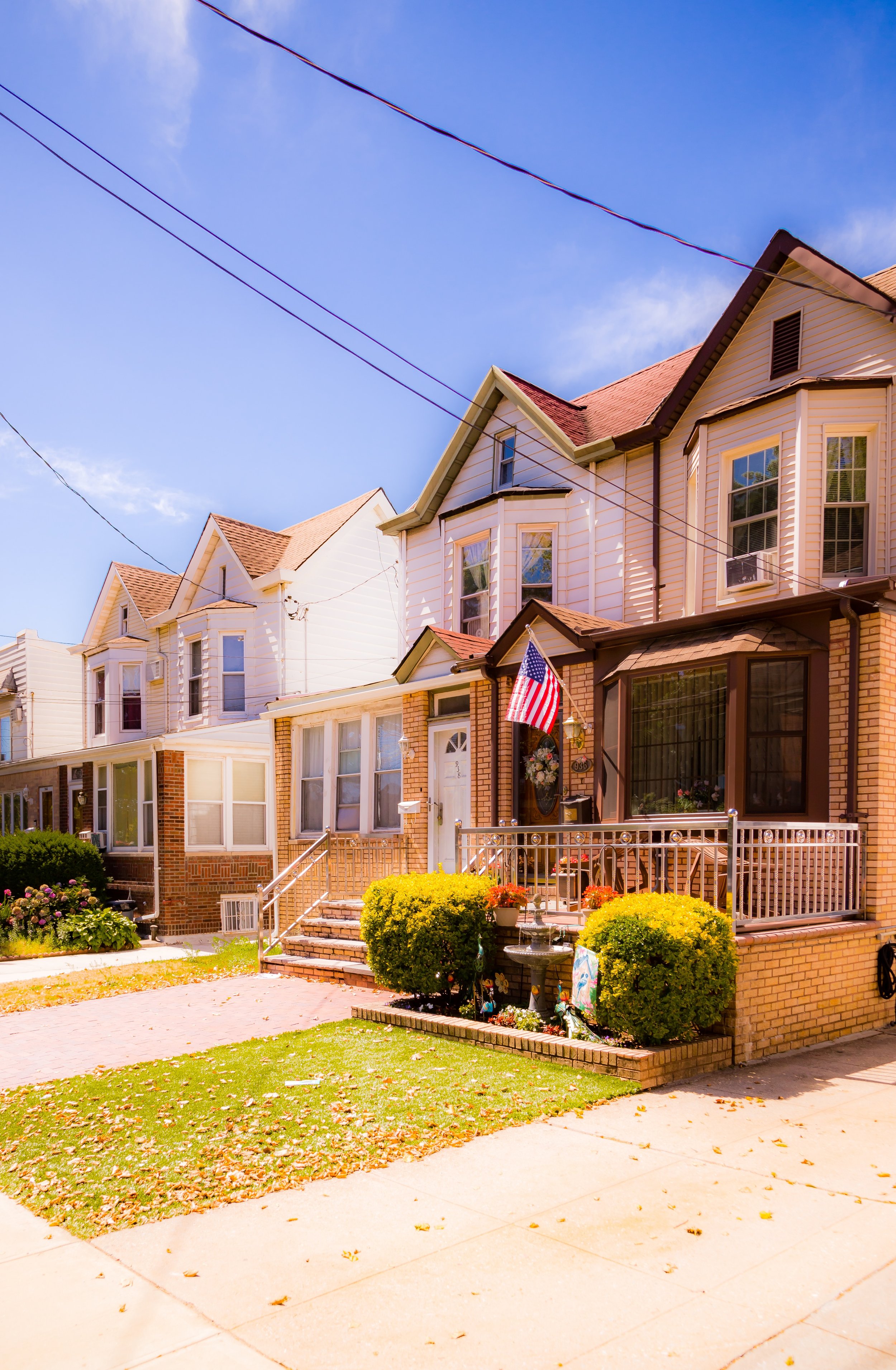 A row of vintage-style houses on a neighborhood street under a bright blue sky, with American flags and front gardens.