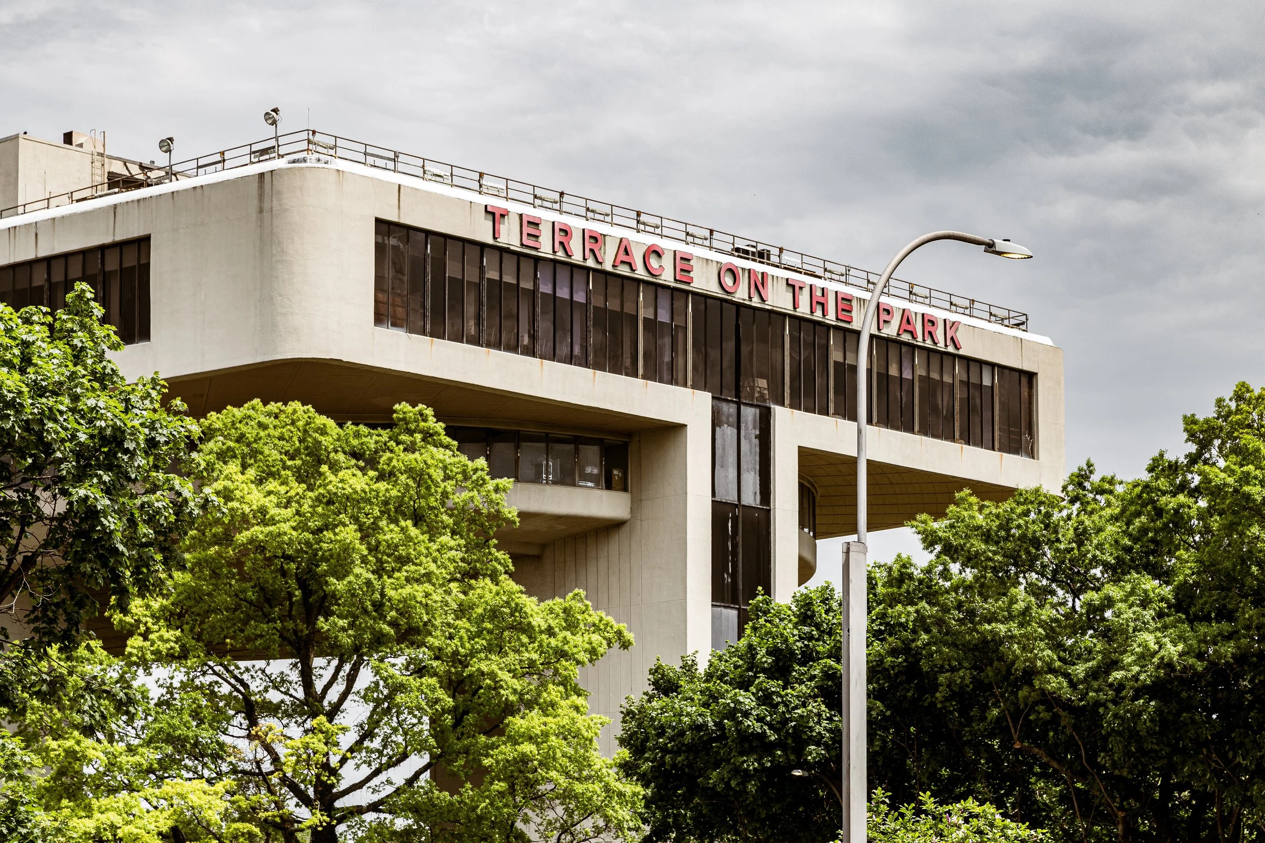 A modern building with large glass windows and a sign that reads 'TERRACE ON THE PARK' on the roof. Green trees are in the foreground, and a street lamp is in front of the building. The sky is cloudy.