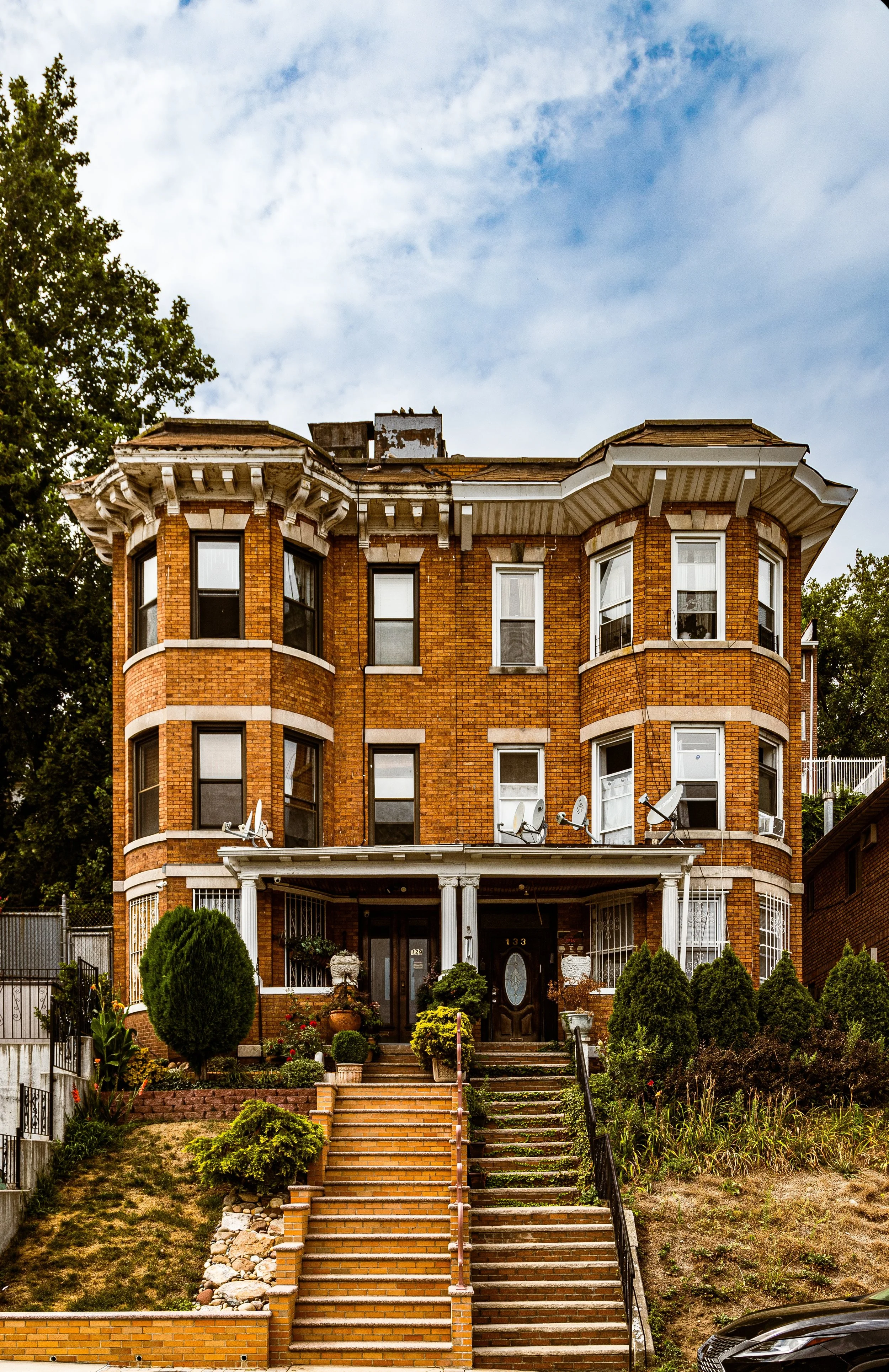 A three-story brick apartment building with a front porch, surrounded by stairs, greenery, and landscaping, set against a partly cloudy sky.