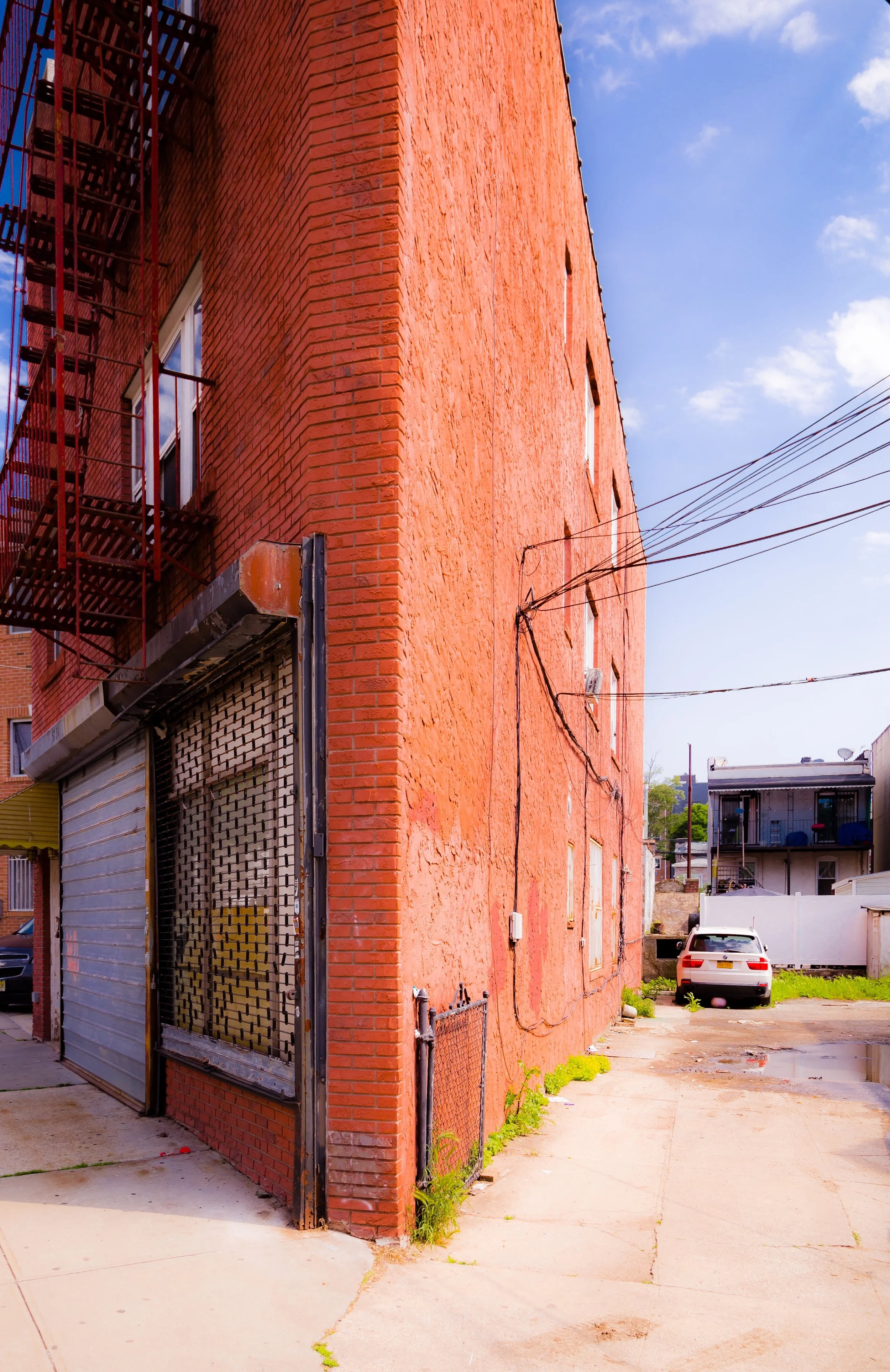 A red brick building with an exterior fire escape, a rolled-down gray metal security gate, and a small fenced area on the sidewalk. A parking lot with a white car is visible in the background under a partly cloudy sky.