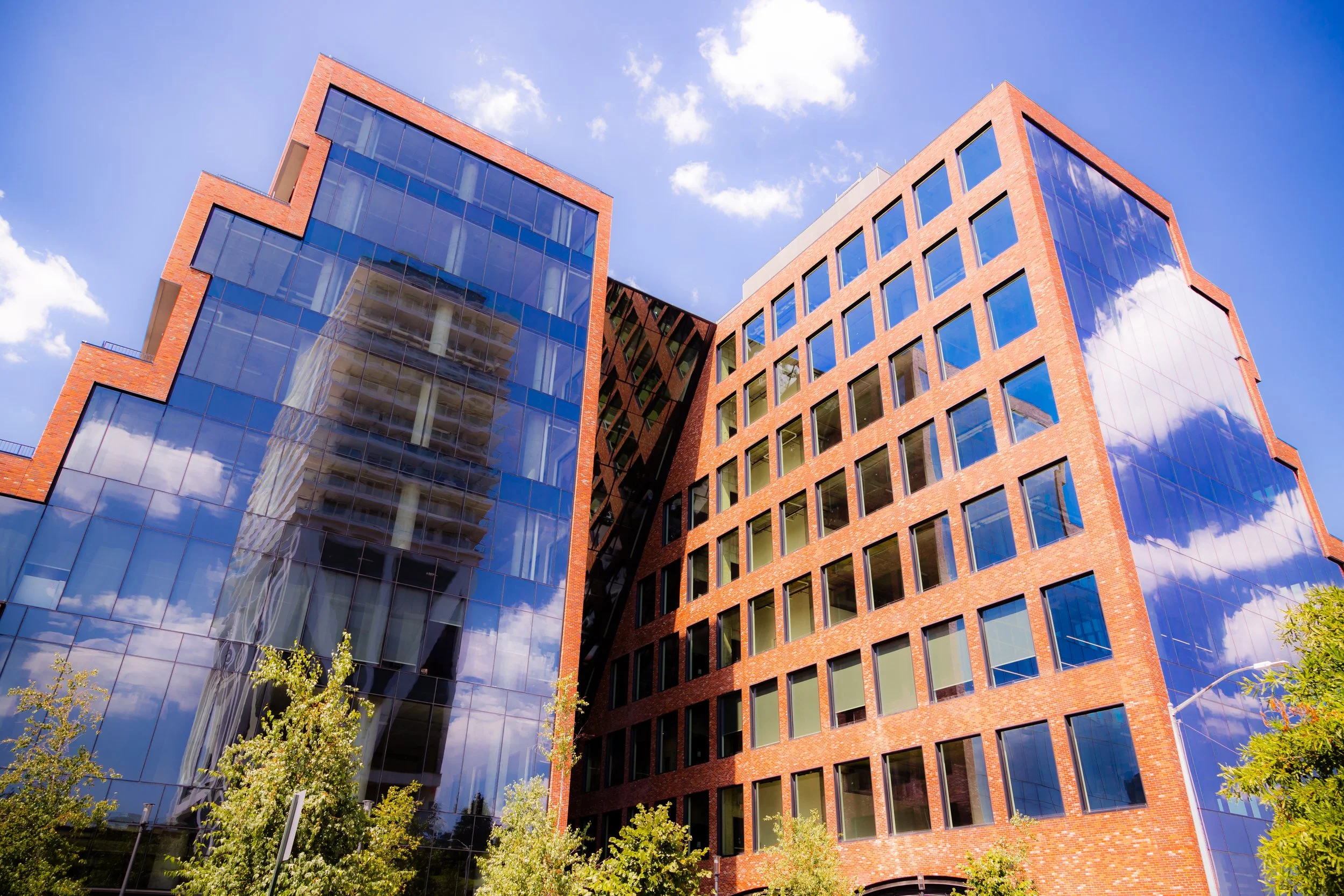 Modern office building with brick and reflective glass facade, under a partly cloudy blue sky with some trees in front.
