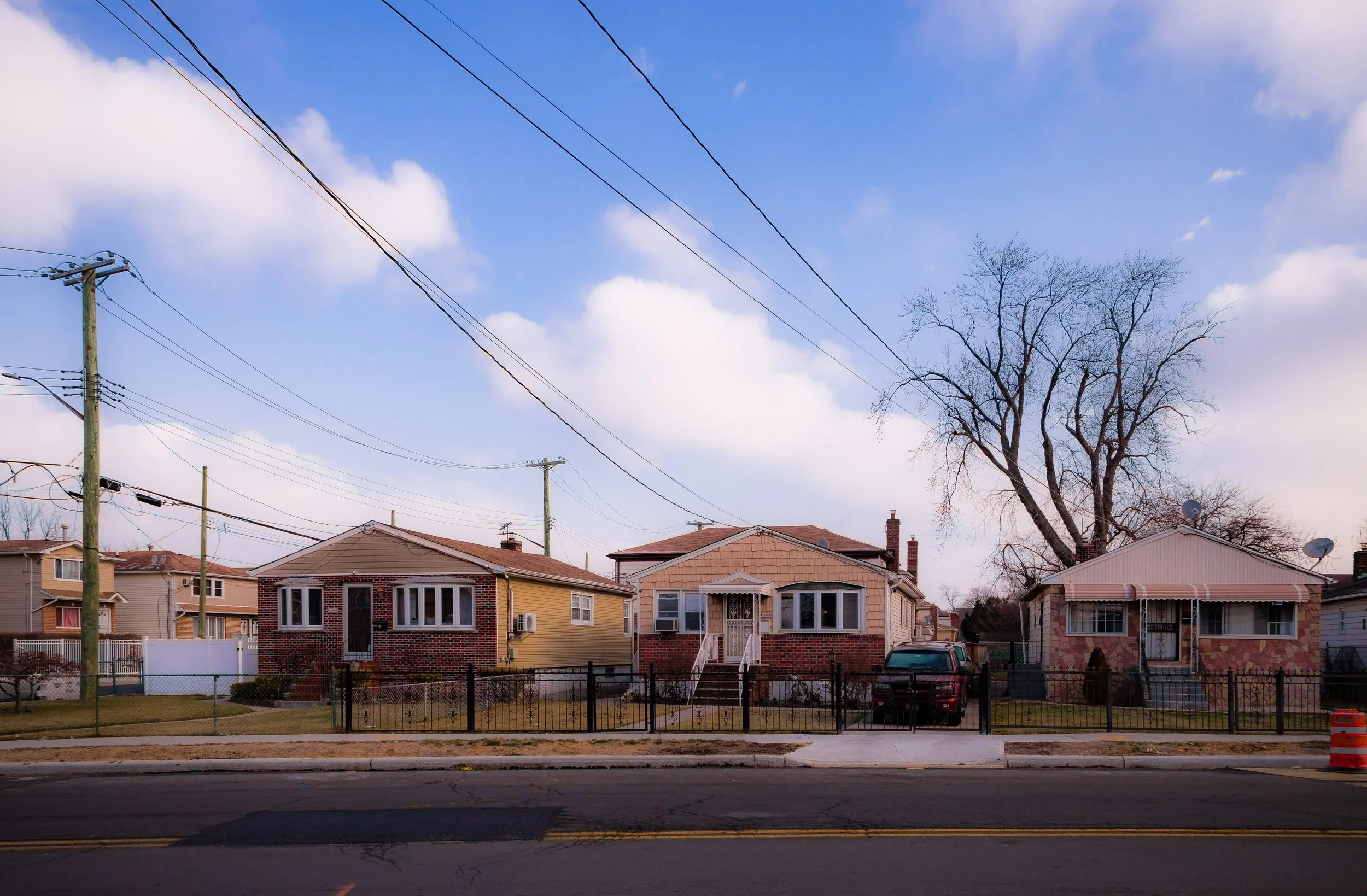 Three single-story houses with small front yards enclosed by fences, a leafless tree, power lines overhead, and a street in the foreground under a partly cloudy sky.