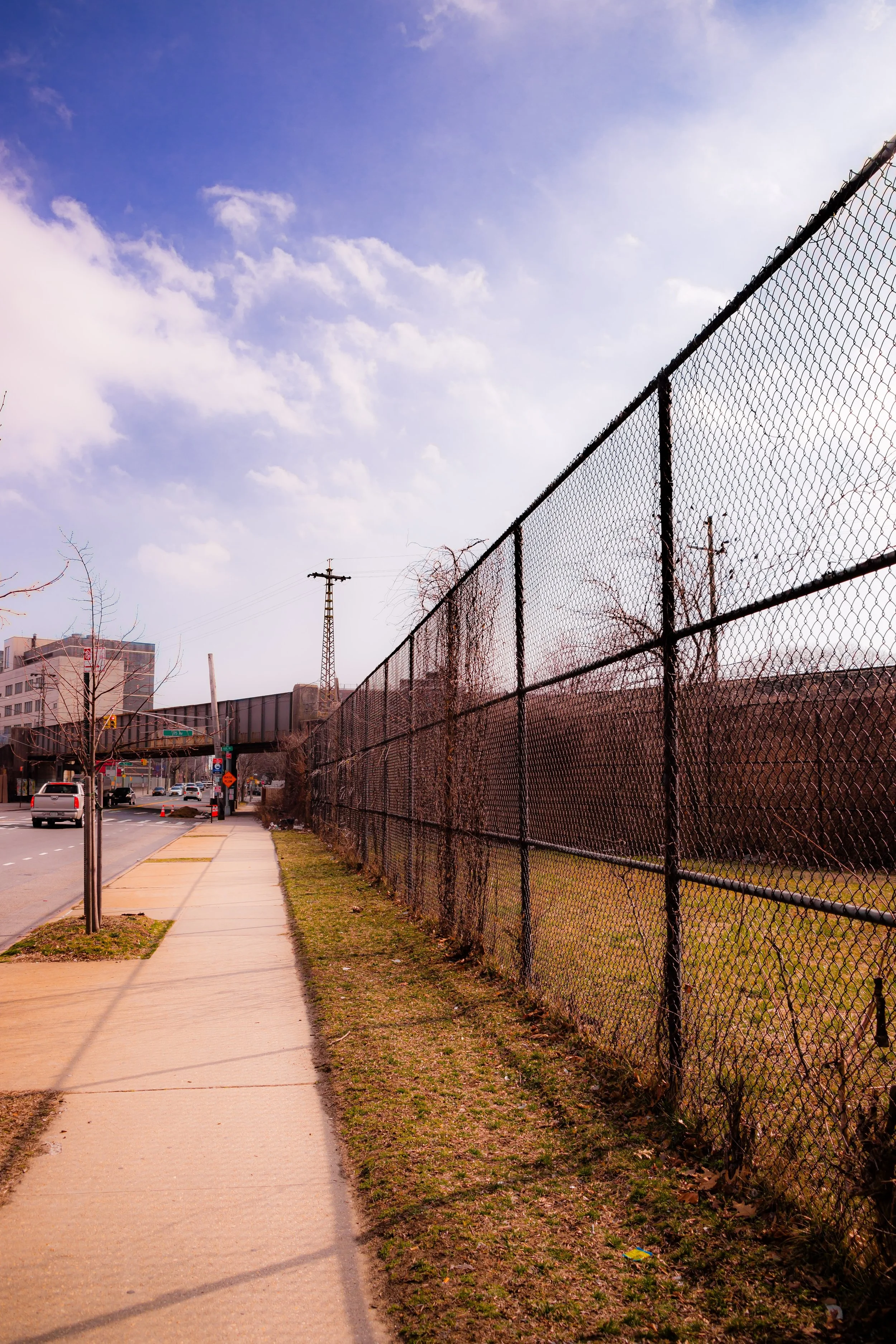 Sidewalk running parallel to a chain-link fence with grass and small trees, with cars and buildings visible in the background on a sunny day with a partly cloudy sky.