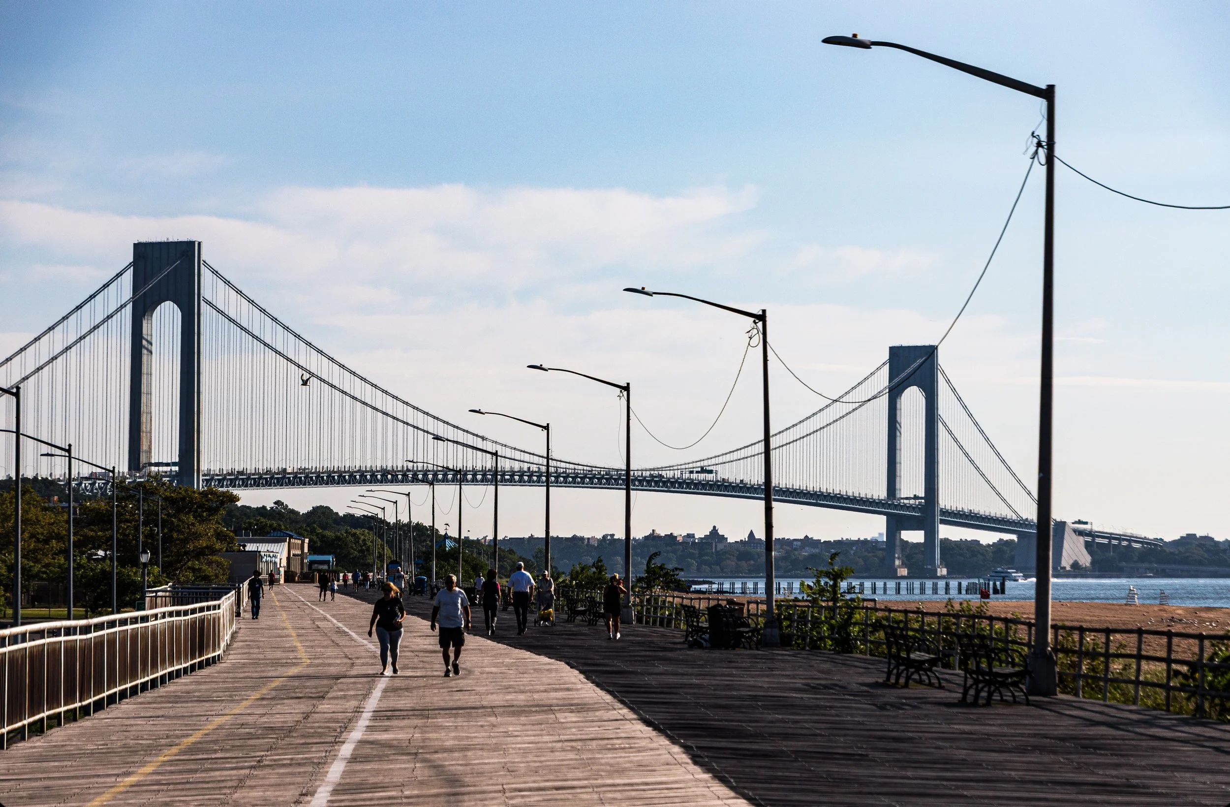 People walking on a wooden promenade with street lamps and benches, with a large suspension bridge spanning a body of water in the background under a partly cloudy sky.