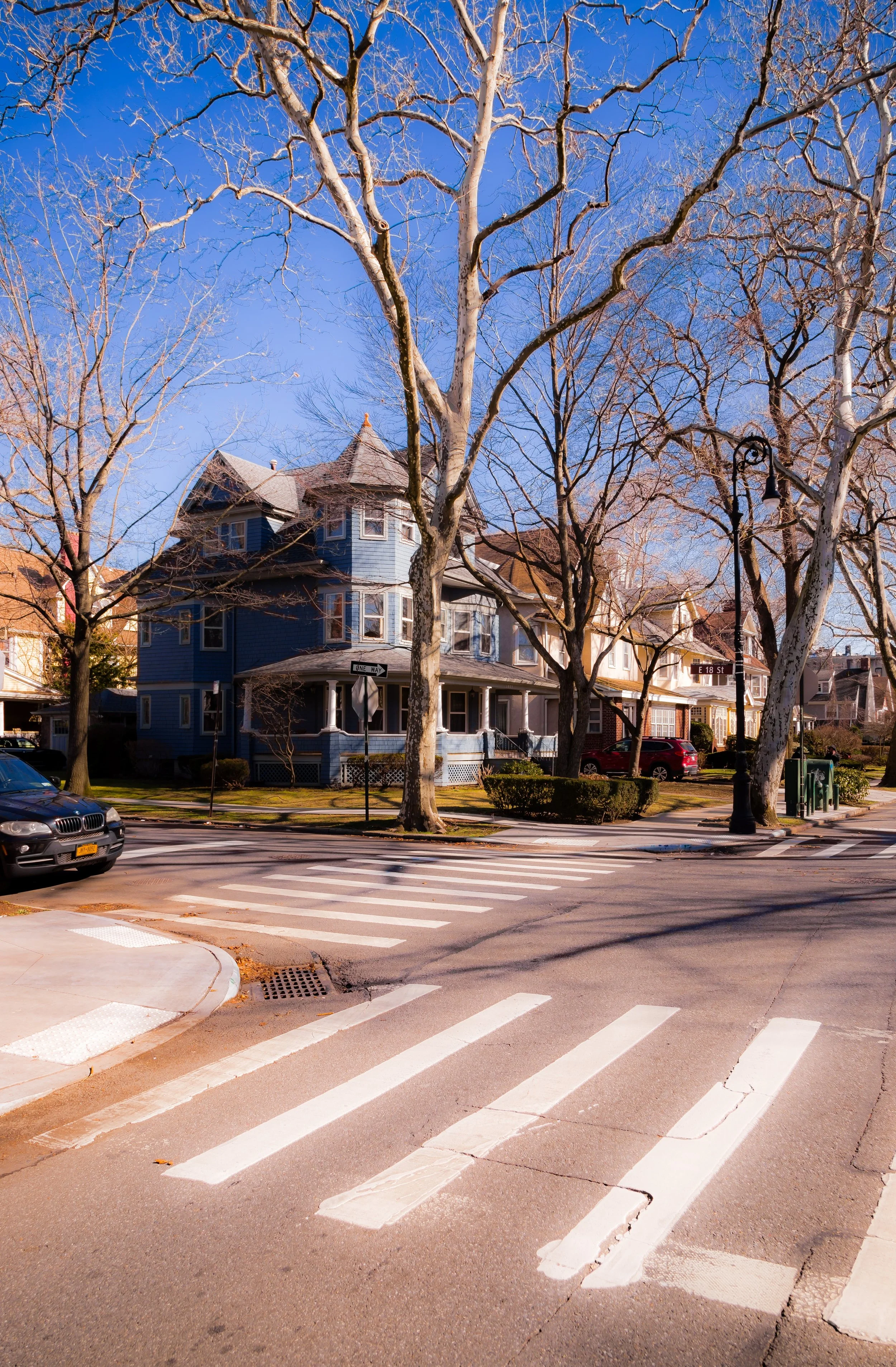 A residential neighborhood scene with a blue Victorian-style house, leafless trees, a crosswalk, a street sign, and parked cars under a clear blue sky.