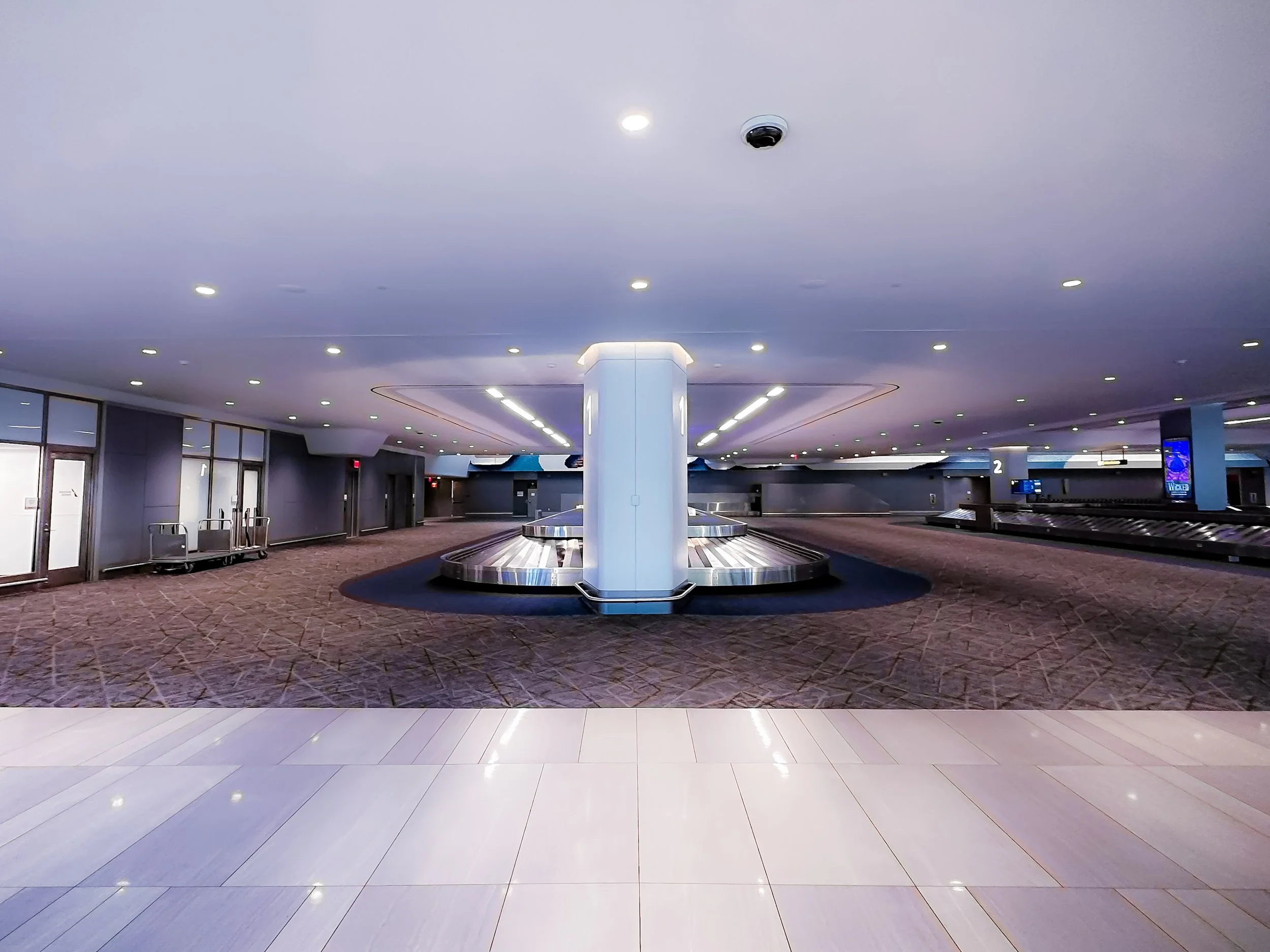 Empty baggage claim area with luggage conveyor belts, a central pillar, and digital screens, in an airport