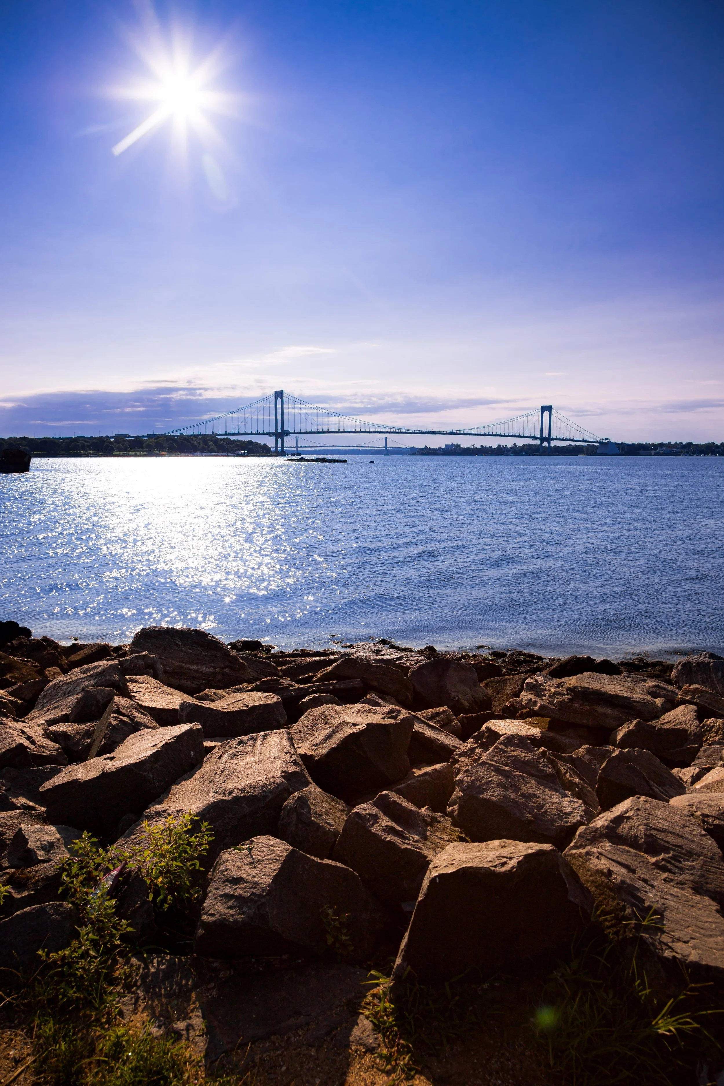 A scenic view of the San Francisco Bay with a bridge in the distance under a clear blue sky, shining sun, rocky shoreline in the foreground, and calm water reflecting sunlight.