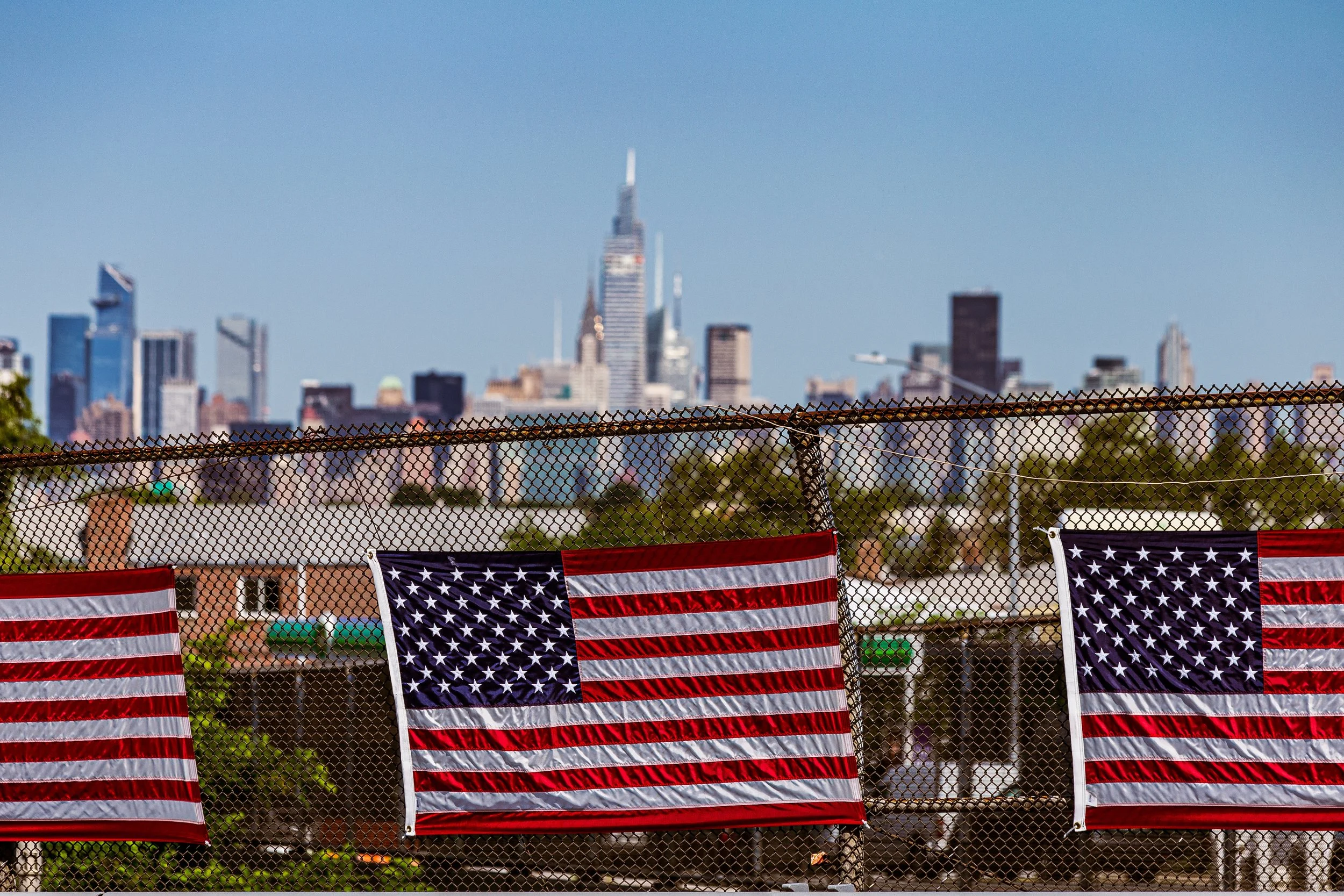 Three American flags hanging on a fence with a city skyline, including the Empire State Building, in the background on a clear day.