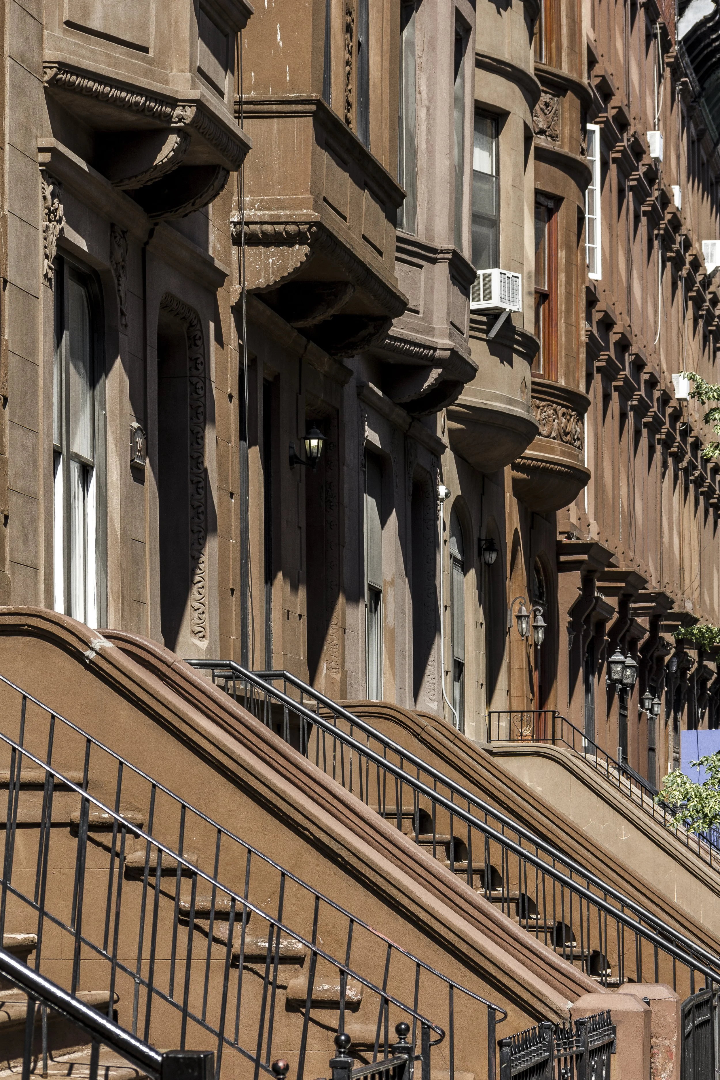 Brownstone row houses with stoops and black railings in an urban neighborhood, featuring ornate architectural details and air conditioning units.