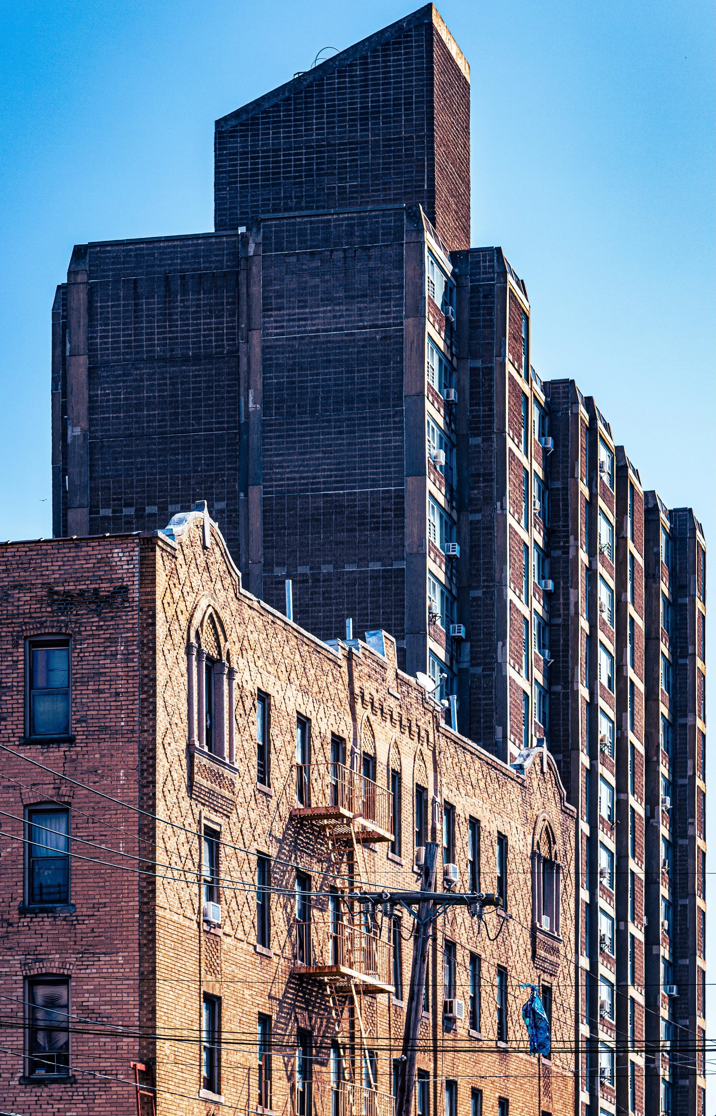 A cityscape featuring a tall, dark modern building behind an older, shorter brick building with fire escape stairs and power lines in foreground.