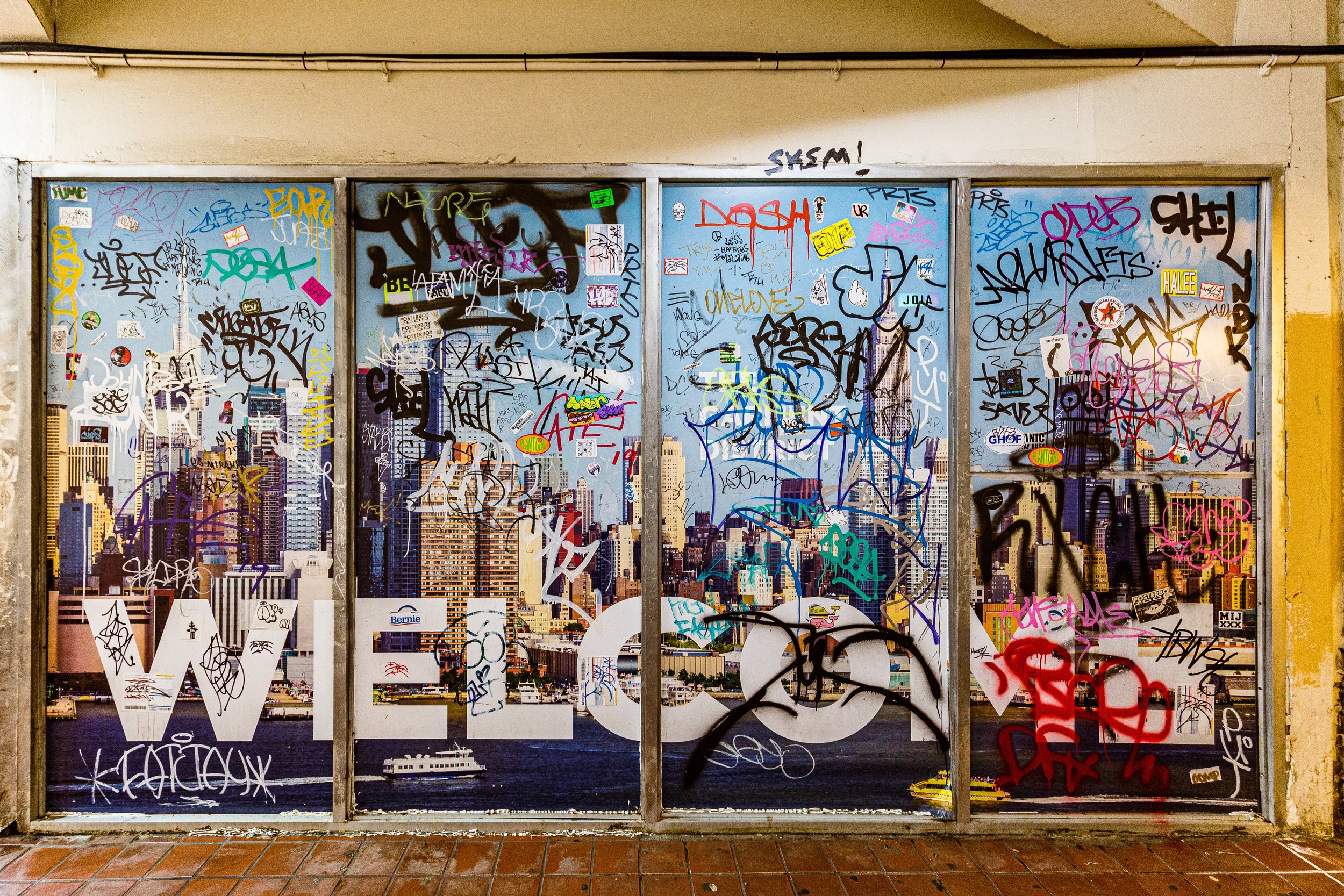 A window with a cityscape view, covered with colorful graffiti tags and stickers.