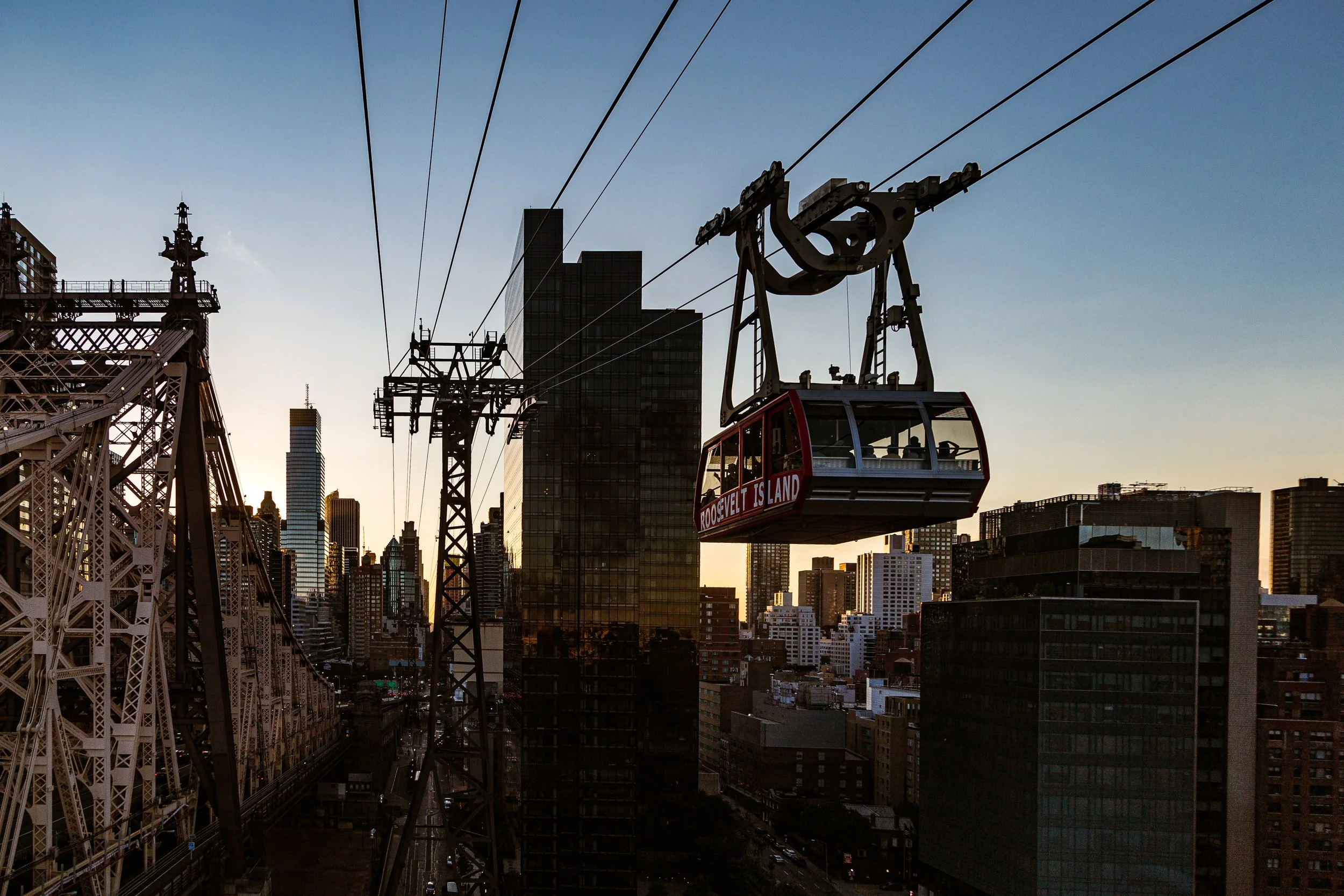 City skyline at sunset with skyscrapers and an observation gondola labeled Roosevelt Island in the foreground.