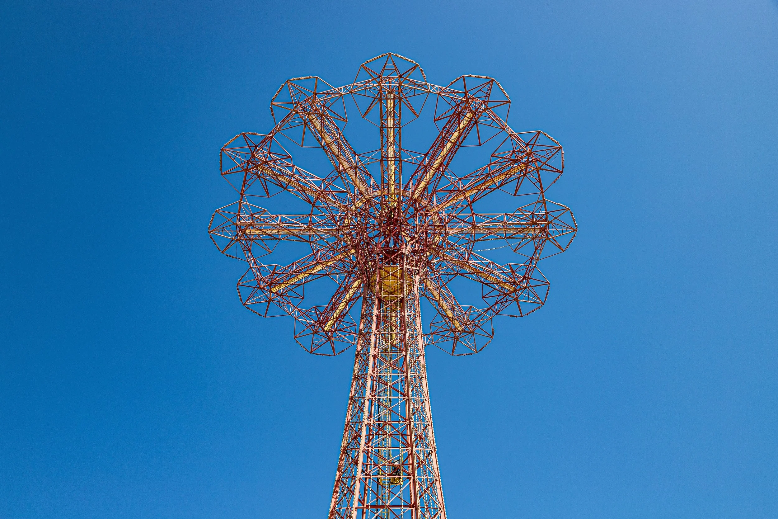 A large, colorful amusement park ride, resembling a Ferris wheel, with angler-style supports and wheel structure against a clear blue sky.