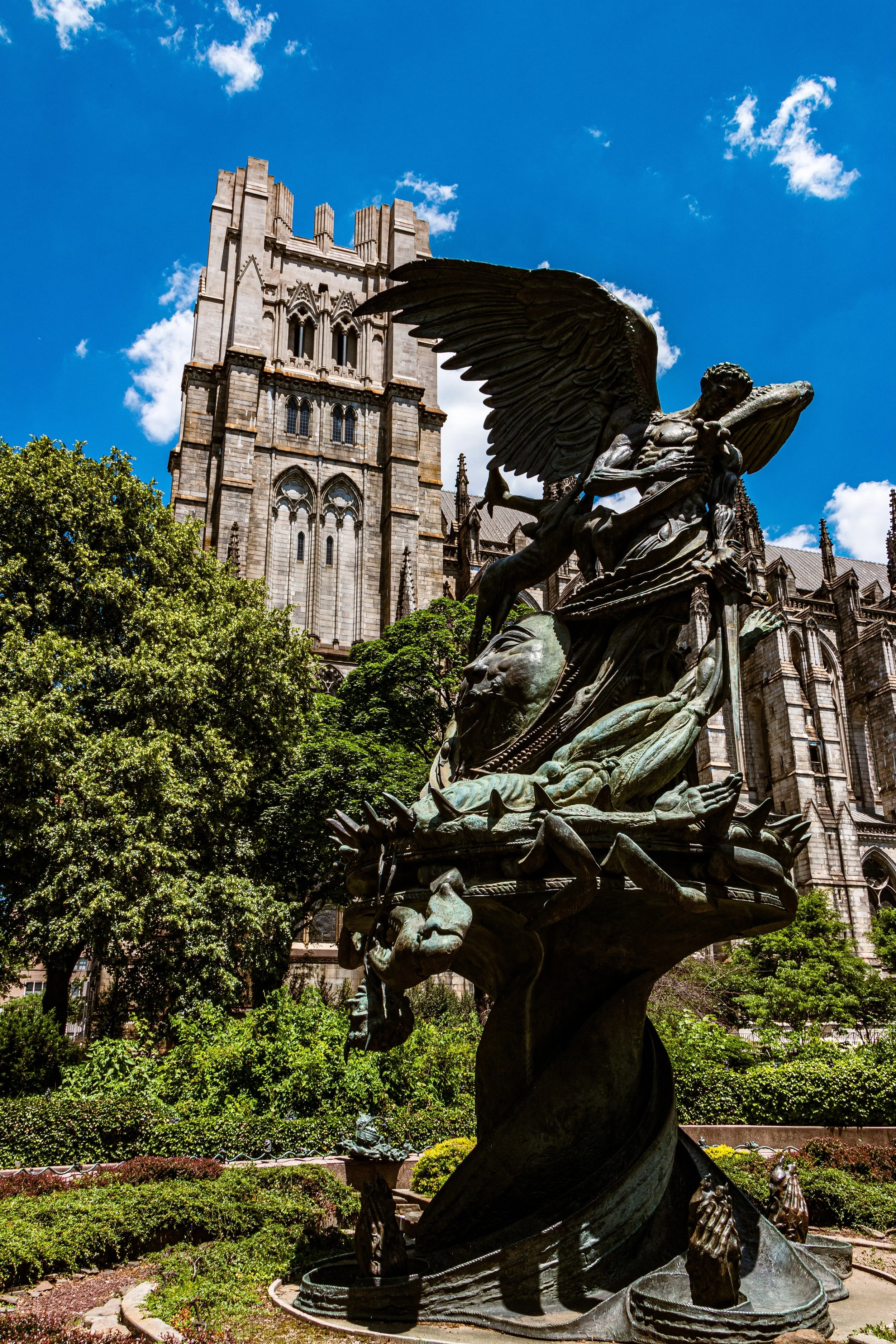 Bronze statue featuring winged figure, lion's head, and mythical creatures in front of a gothic cathedral with blue sky and green trees.