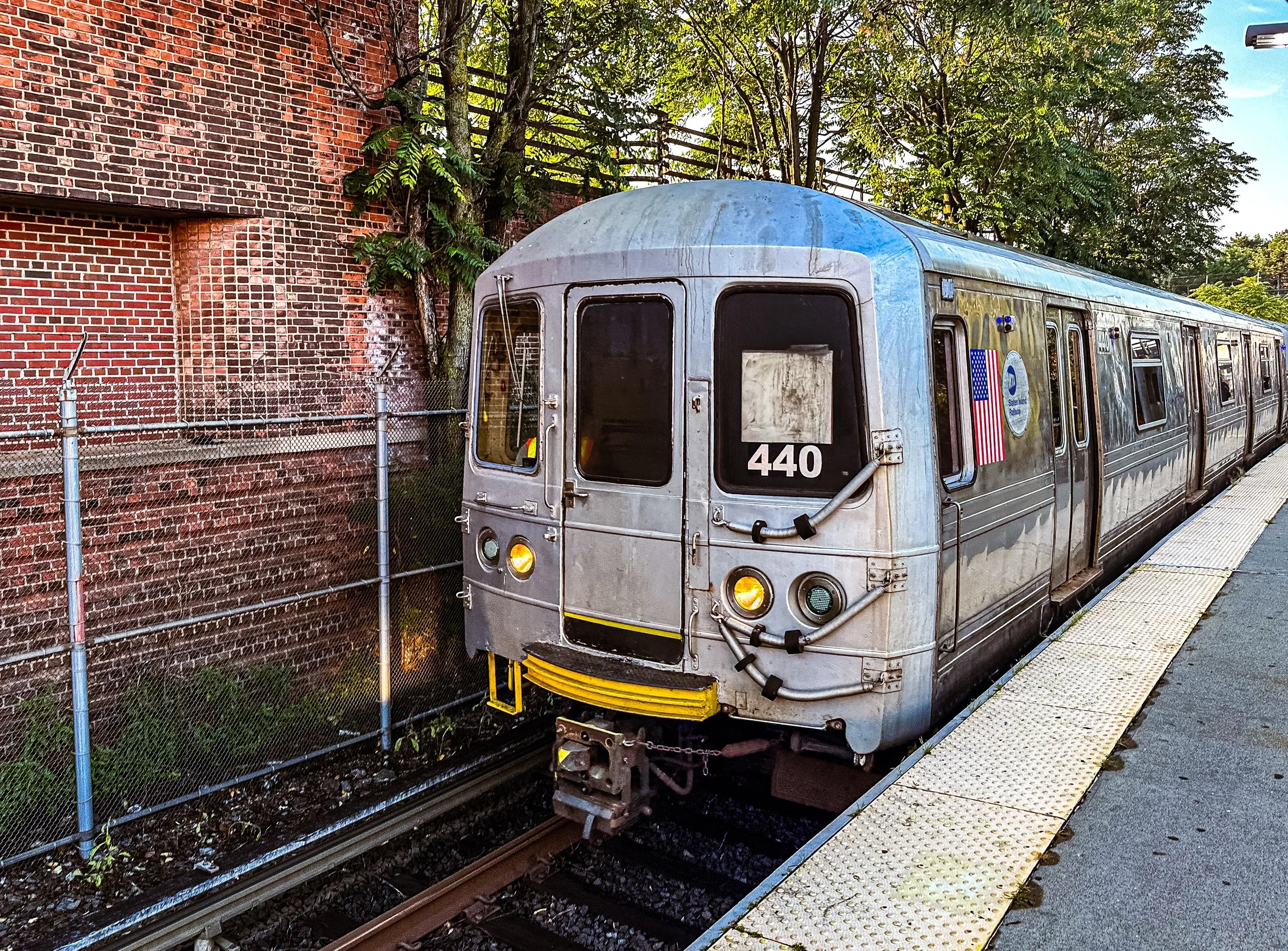 An urban train stopped at a station platform, with a brick wall and greenery in the background. The train is metallic silver with yellow accents and has the number 440 on its front.