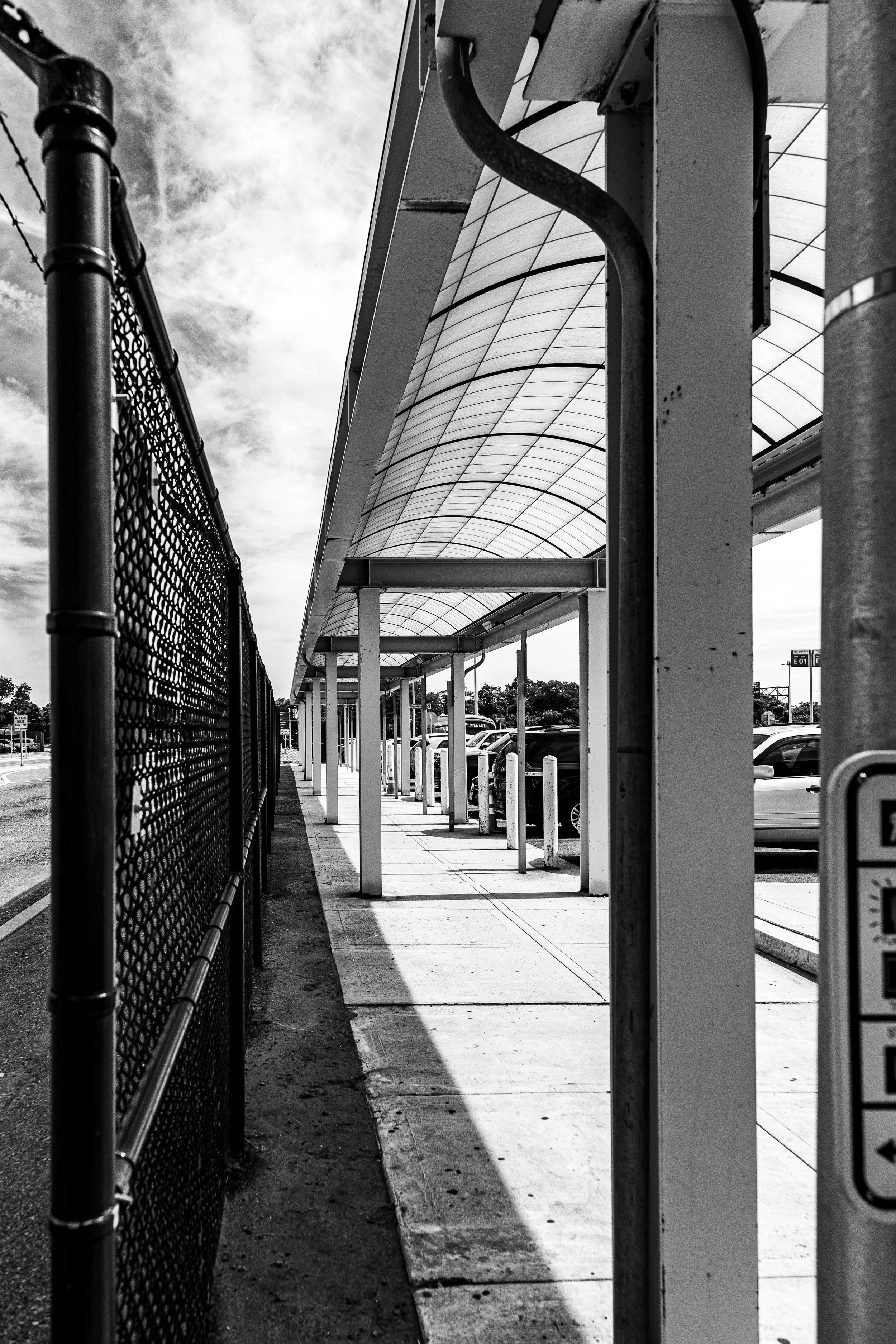 A black-and-white photo of a parking lot with a covered walkway, parked cars, a fence on the left, and a partly cloudy sky.