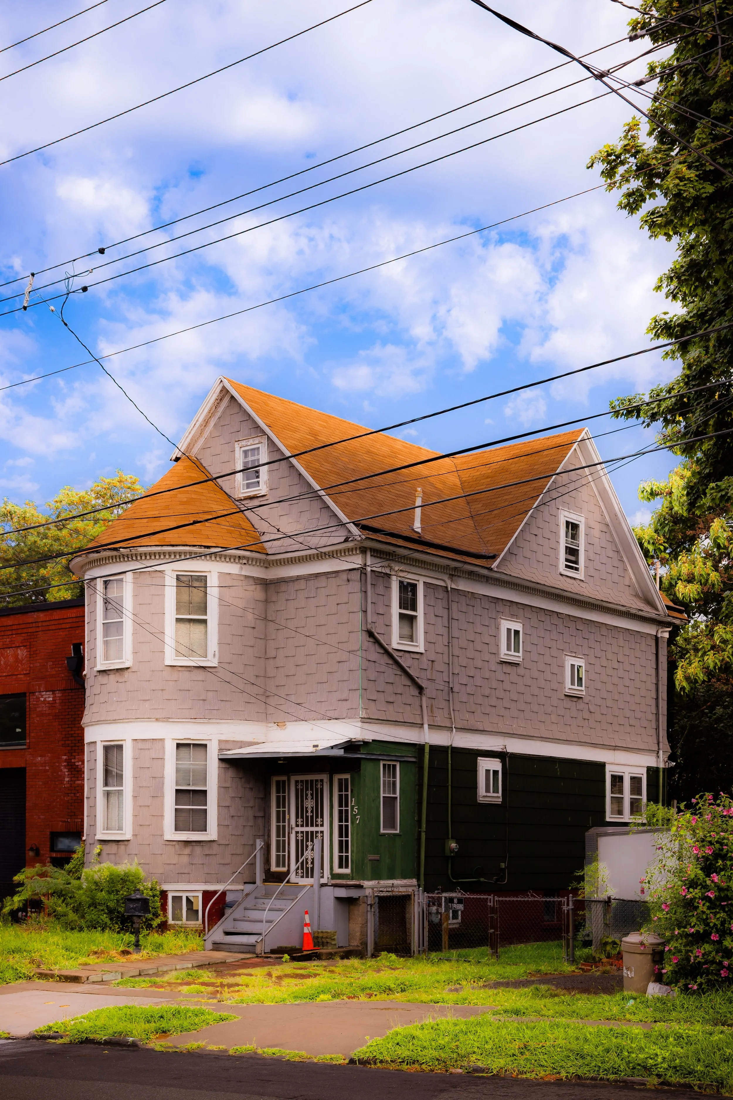 A three-story house with a rounded turret, tan exterior shingles, and an orange roof, situated in a residential neighborhood with trees and a cloudy sky.