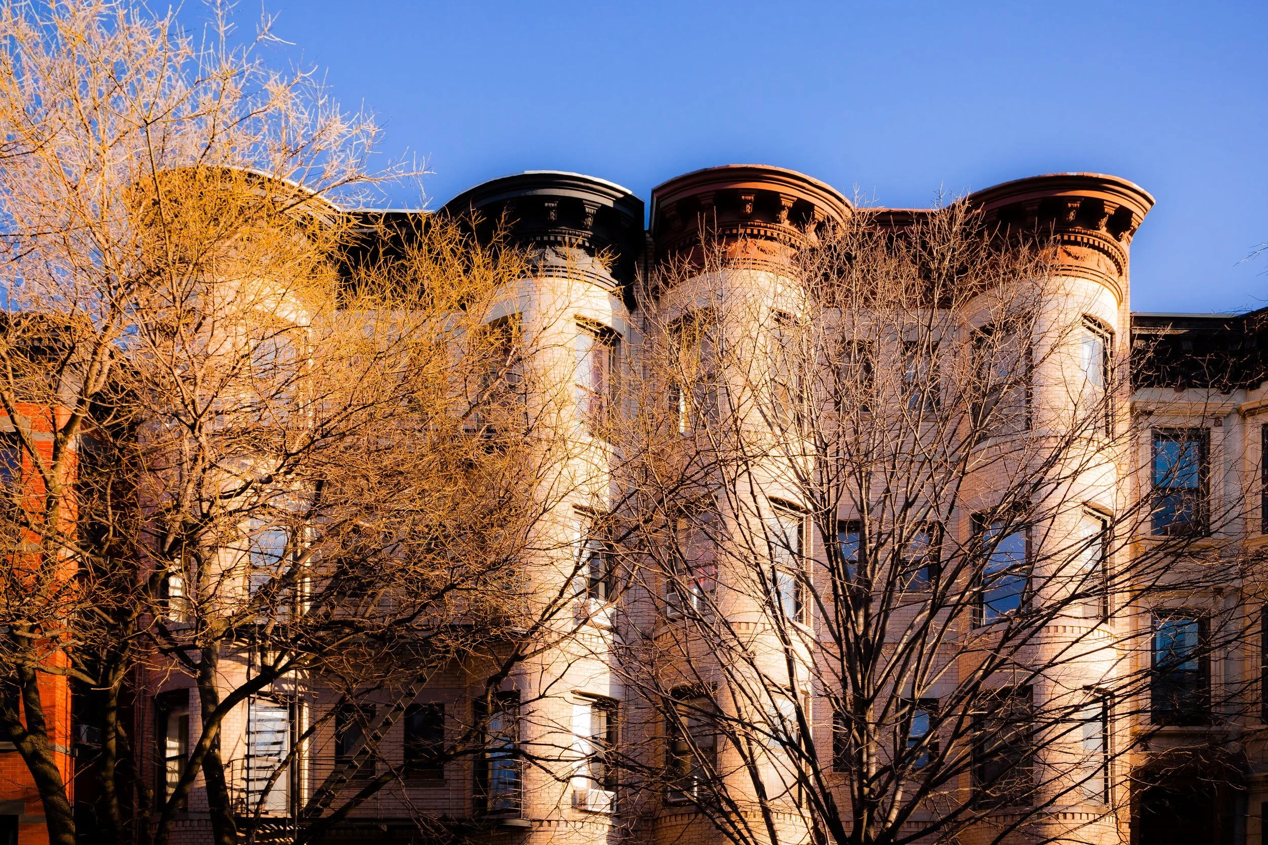 Brownstone city building with leafless trees in front, under a clear blue sky during sunset.