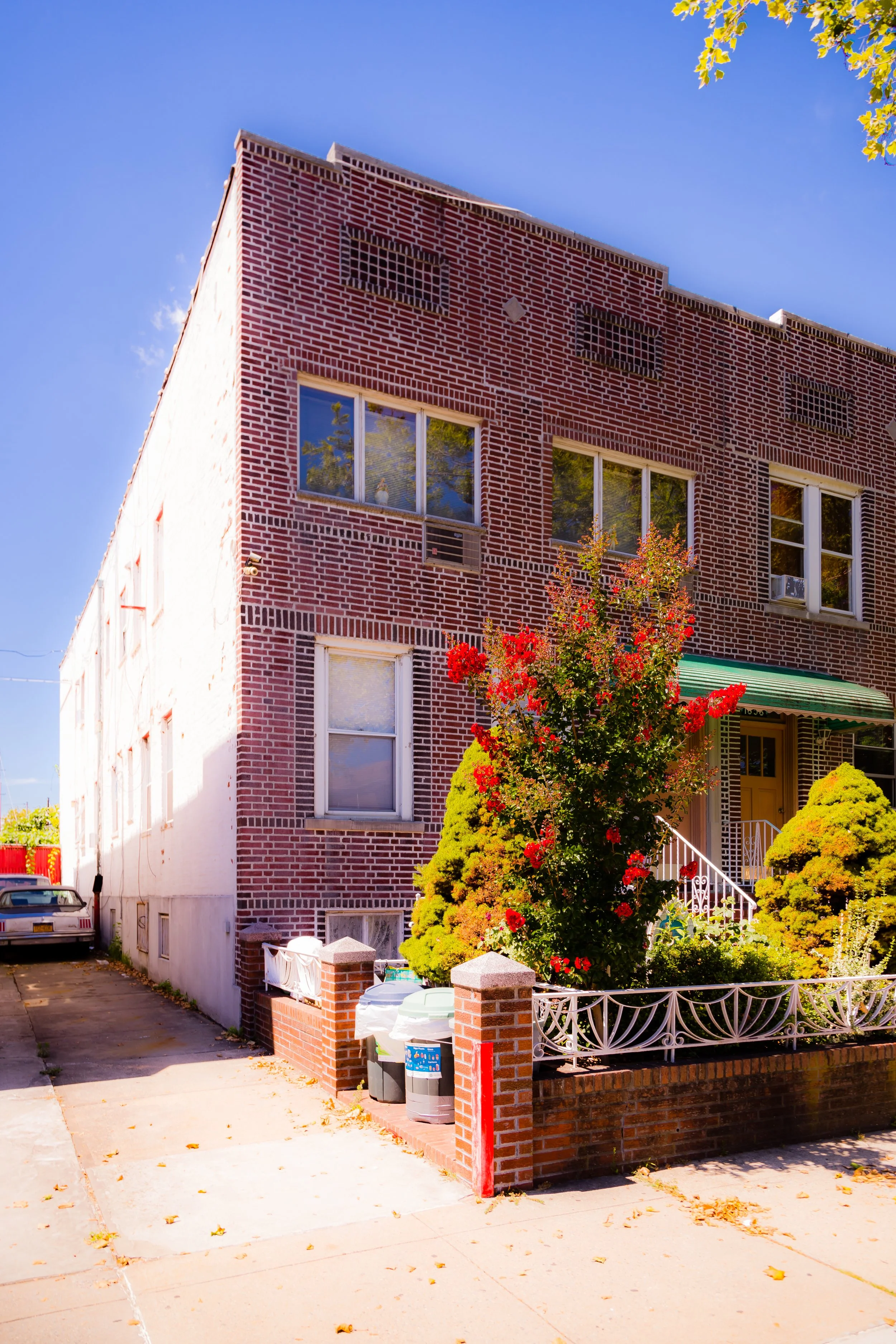 A three-story brick residential building with multiple windows and a green awning over the entrance. There is a small garden with bushes and a tree with red flowers in front, and a sidewalk and driveway with a car parked beside the house. The sky is clear and blue.