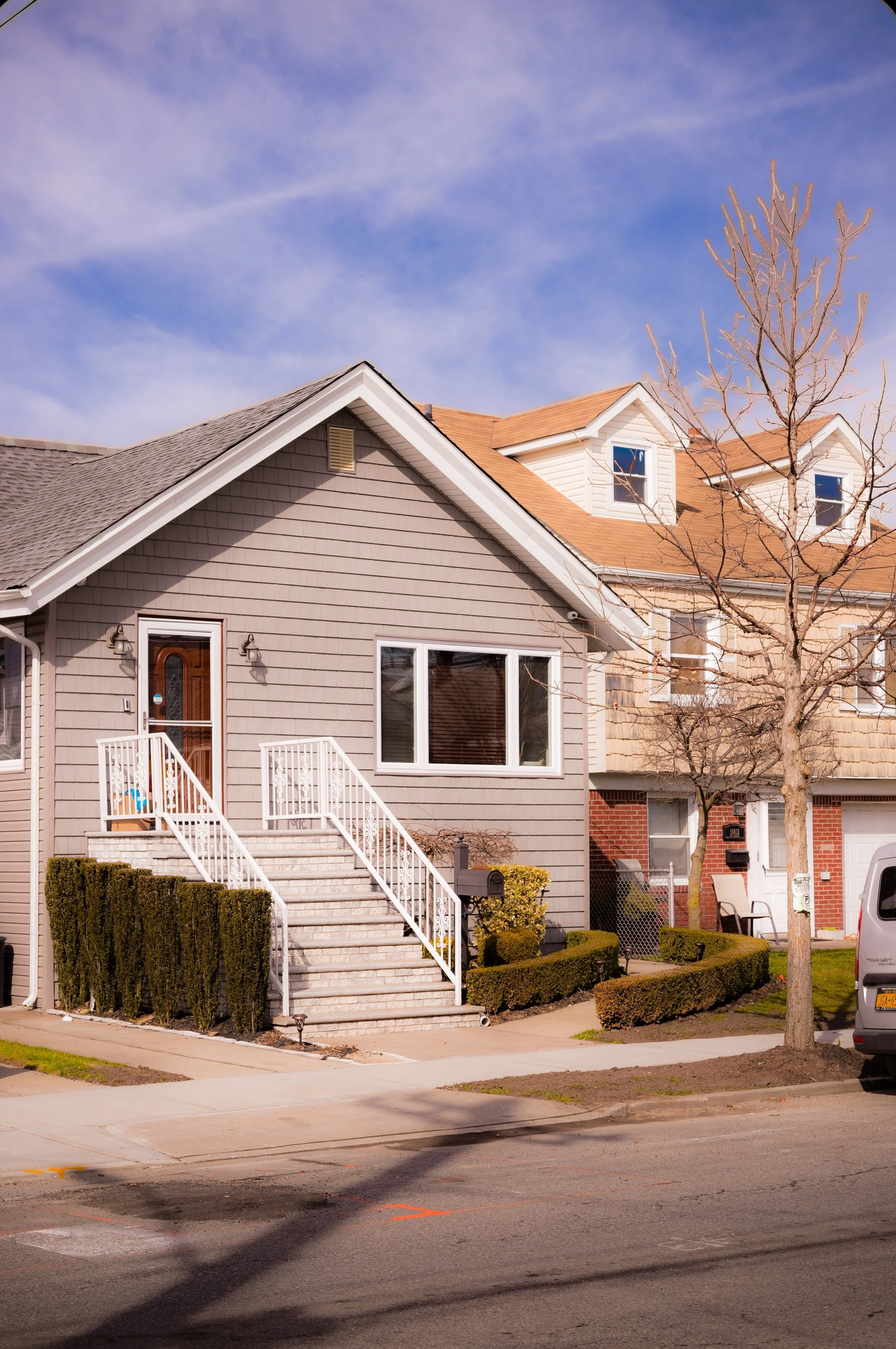 A suburban house with grey siding, white stairs, and beige roof, with a leafless tree and a parked vehicle visible in front.