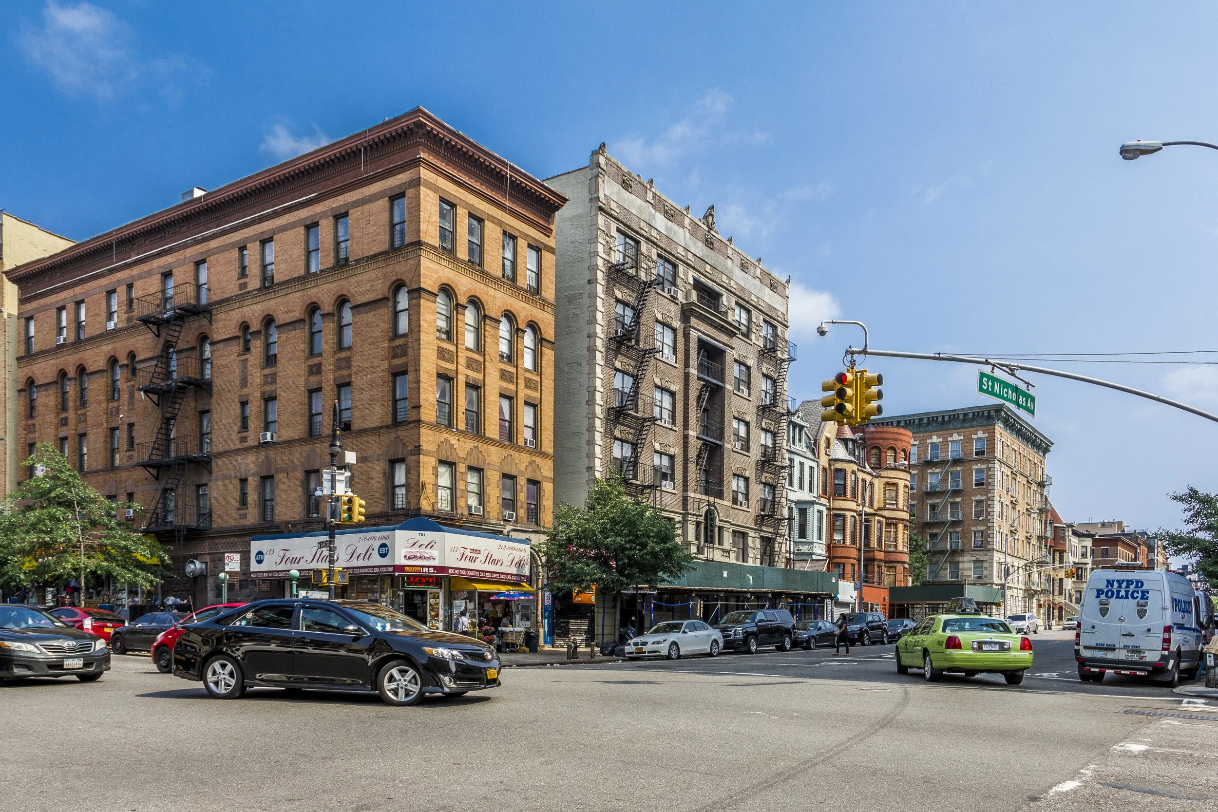 City street scene with brick apartment buildings, cars parked along the street, a police van, traffic lights, and a street sign reading 'St. Nicholas Ave' under a blue sky.