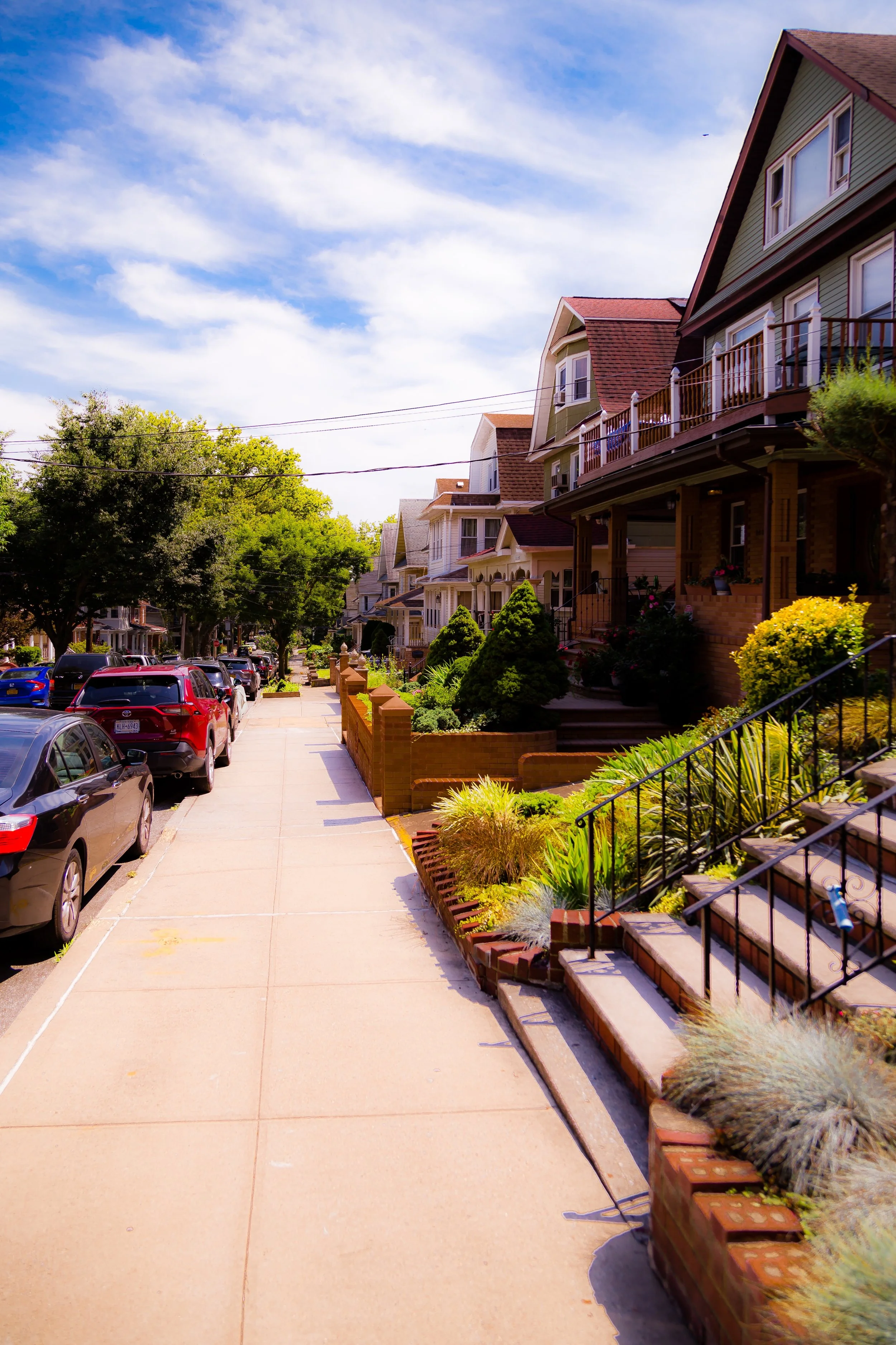 A sunny residential street with parked cars on the left, lined with colorful houses and well-maintained front yards on the right, and a blue sky with some clouds overhead.