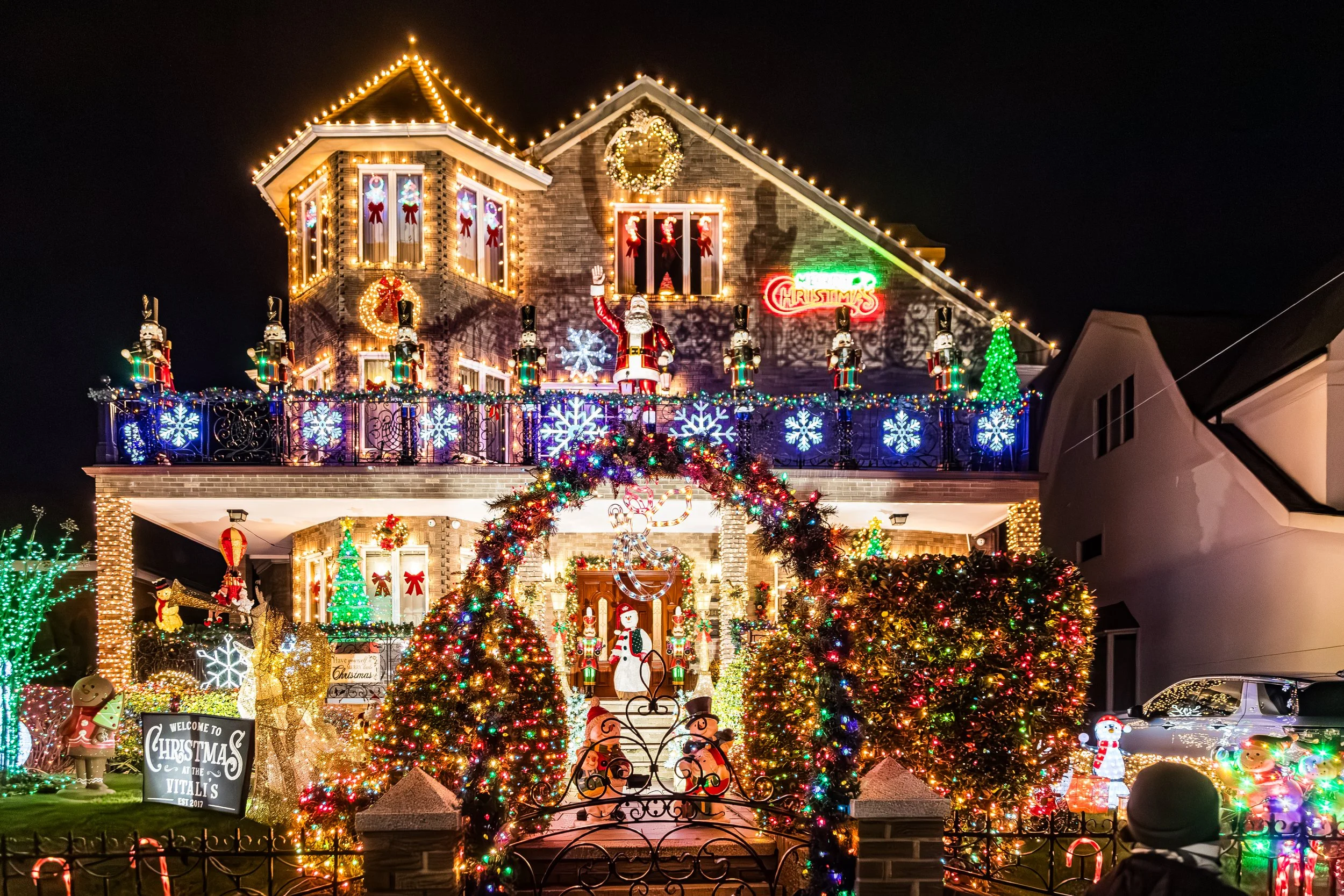 Festively decorated house with Christmas lights, inflatables, and ornaments at night, including snowflakes, a snowman, Santa, reindeer, and a sign that reads 'Welcome to Christmas at the Vitali's'.