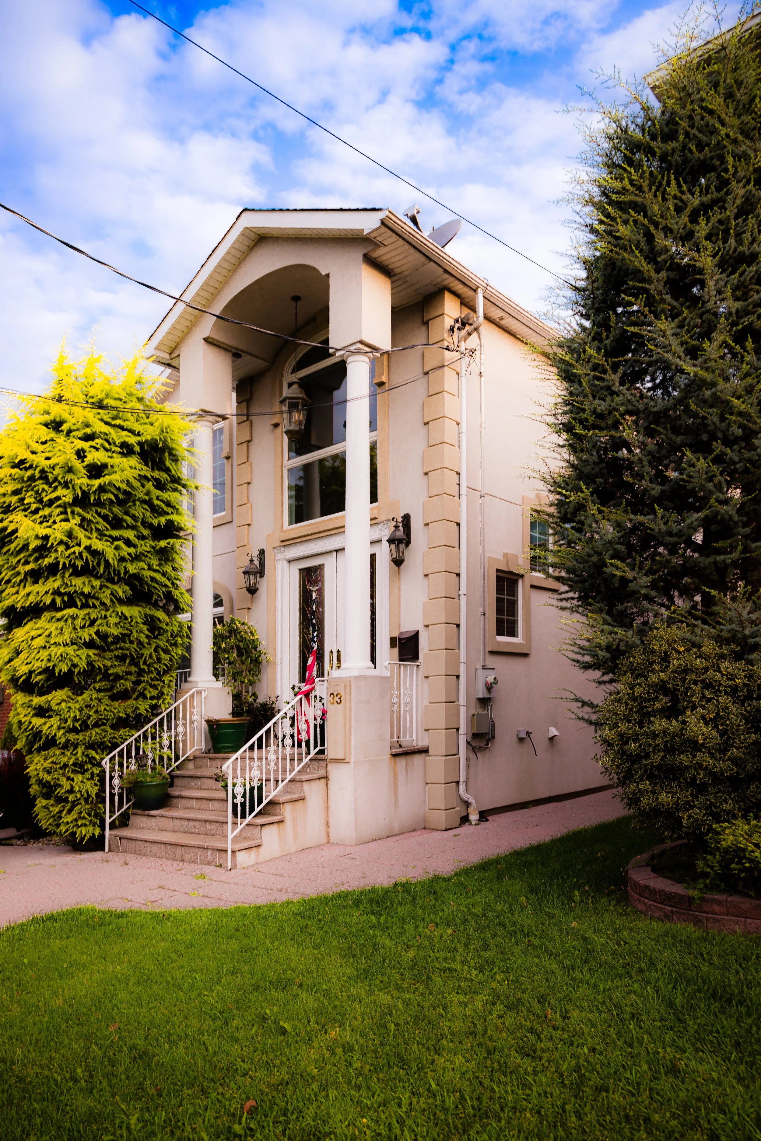 A two-story house with a front porch, white columns, and large windows, surrounded by green lawn and tall trees, under a partly cloudy sky.