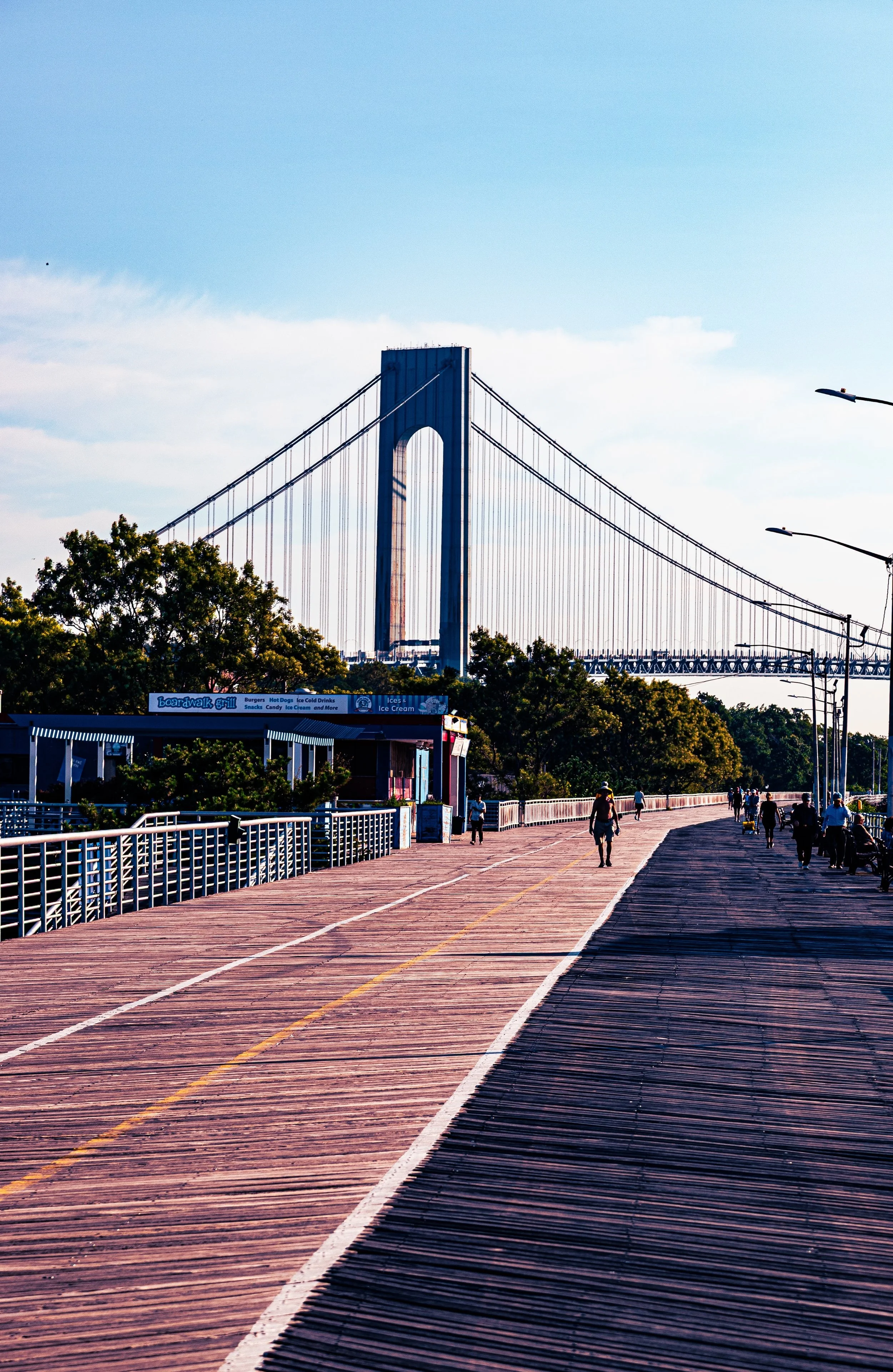 A wooden boardwalk with people walking, and the Brooklyn Bridge visible in the background under a blue sky with some clouds.