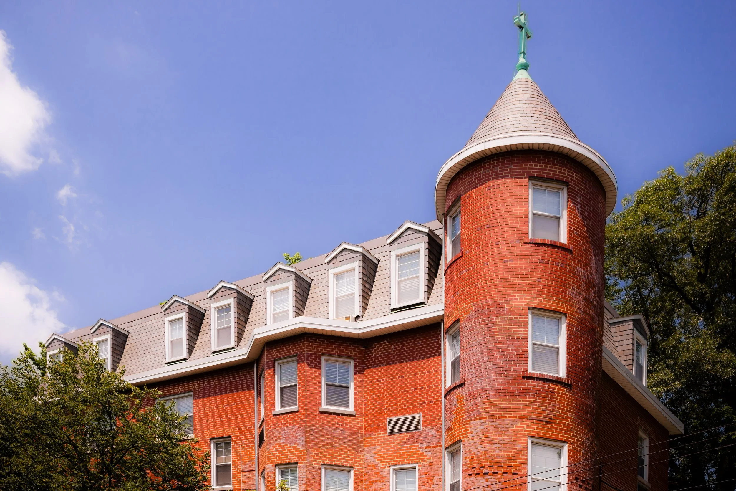 A historic red brick apartment building with a distinctive rounded turret topped with a cross and several dormer windows, set against a partly cloudy sky and surrounded by trees.