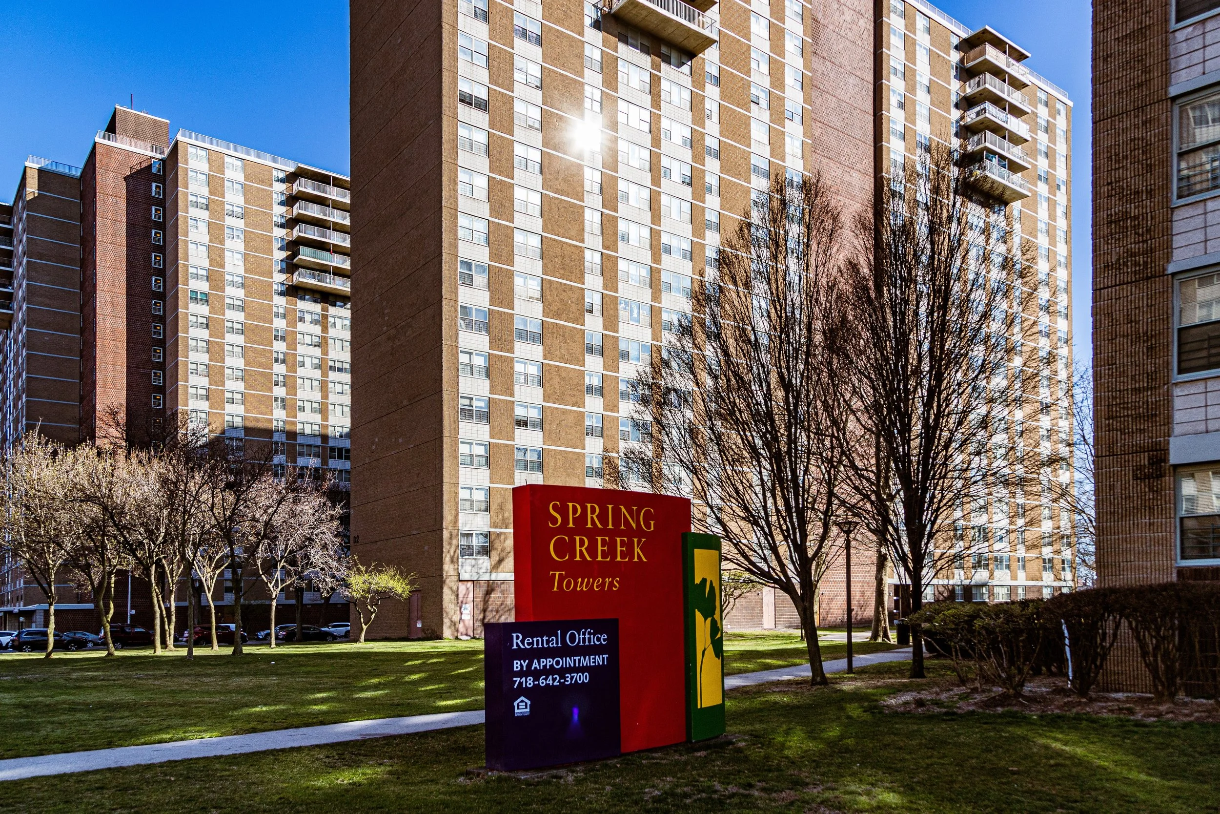 Apartment complex with tall, multi-story buildings, a grassy area, leafless trees, and a red and yellow sign that reads "Spring Creek Towers." The sign also indicates rental office information with a phone number and a small house icon.