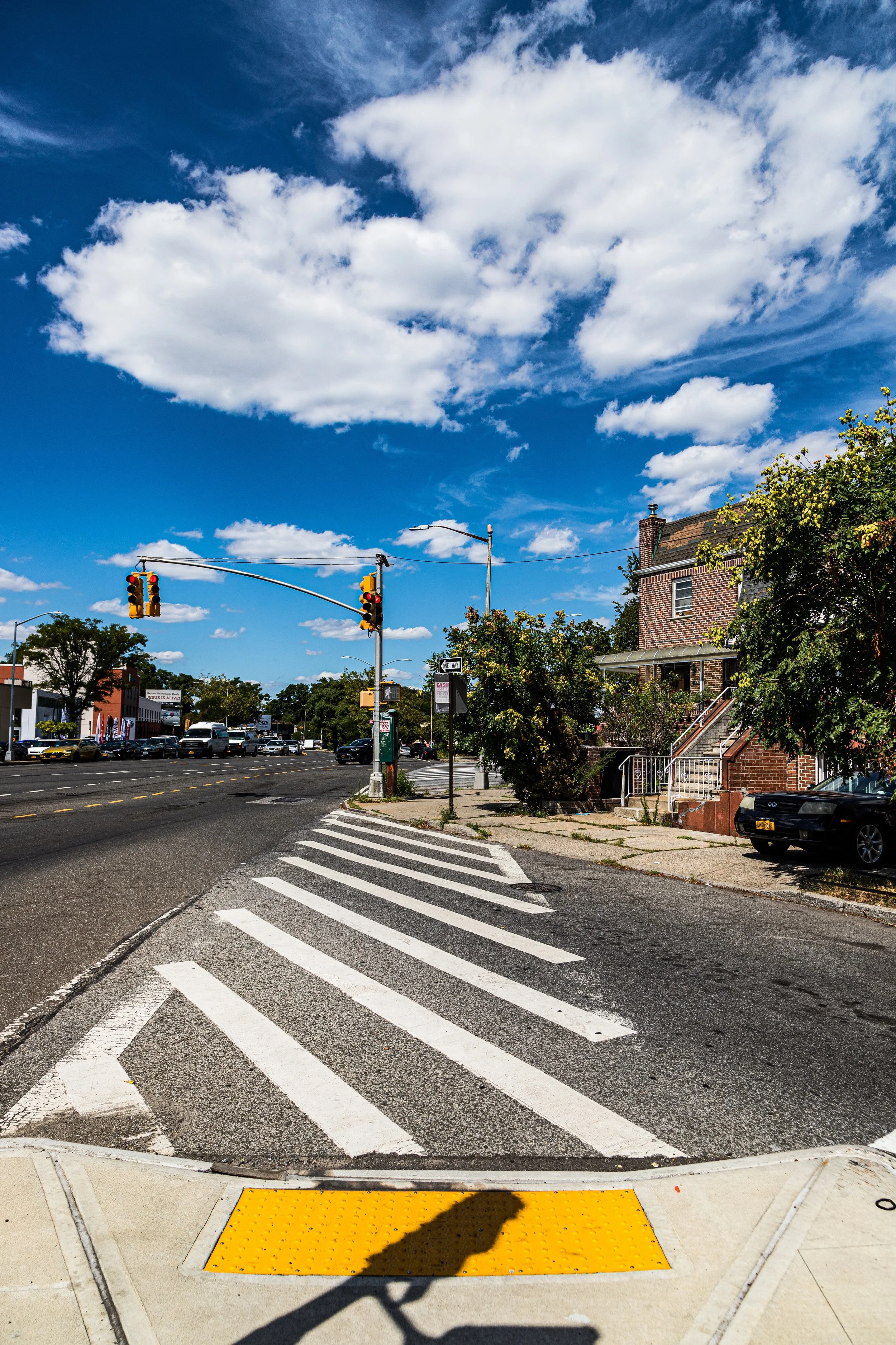 A city street intersection with traffic lights, crosswalk markings, and parked cars under a partly cloudy blue sky.